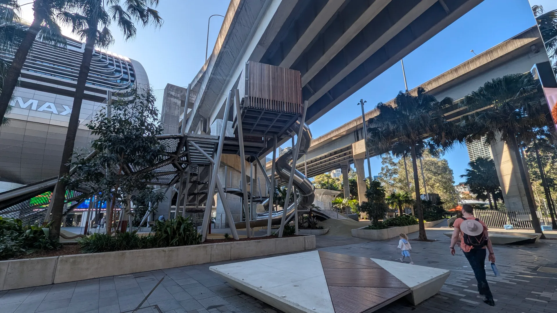 A modern, multi-level metal playground with a spiral slide and climbing nets, set beneath an urban highway overpass with palm trees and paved paths.