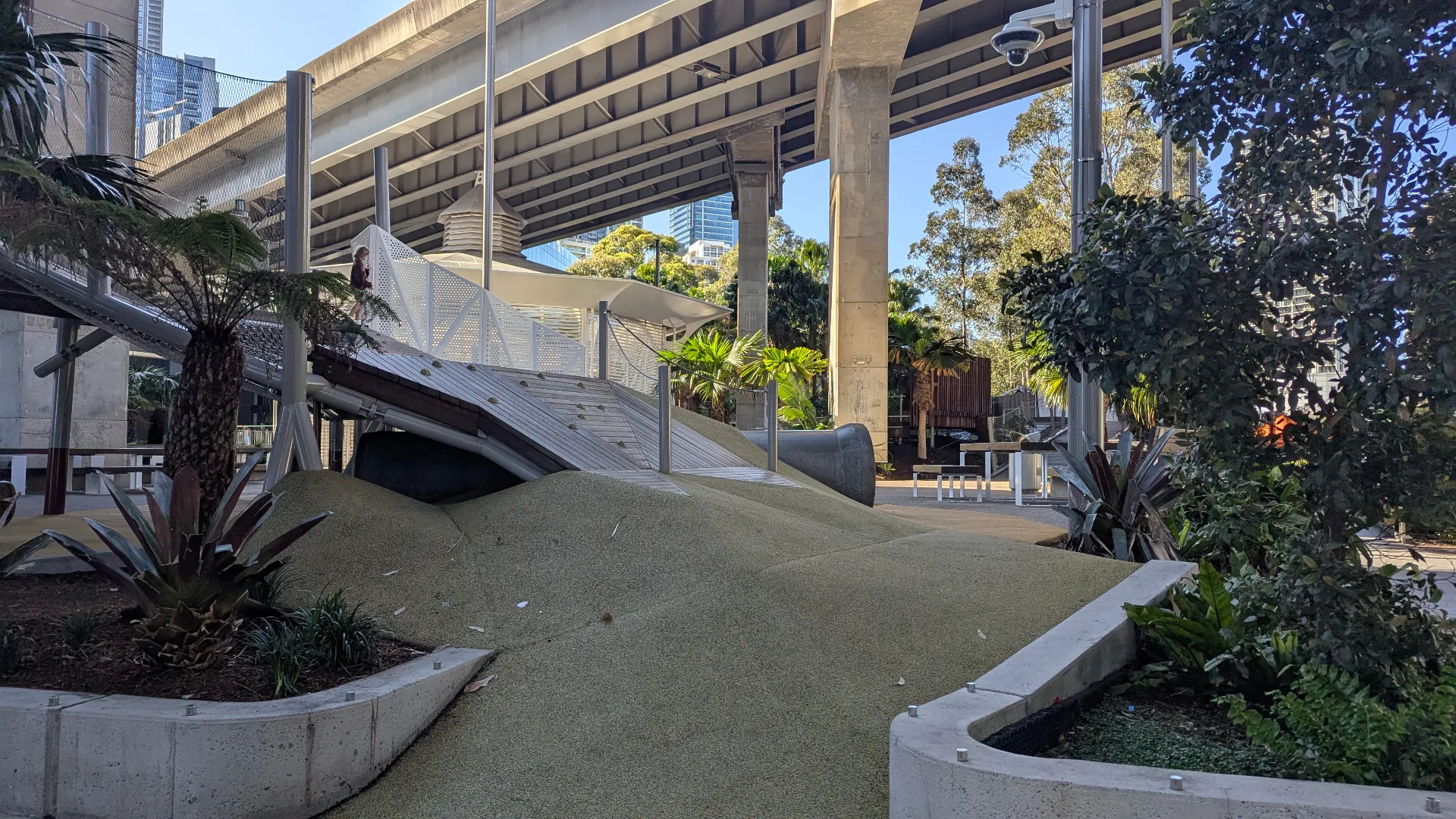 A sloped climbing ramp and rubber-surfaced mound in an urban playground, featuring wooden slats, handrails, and surrounding tropical greenery.