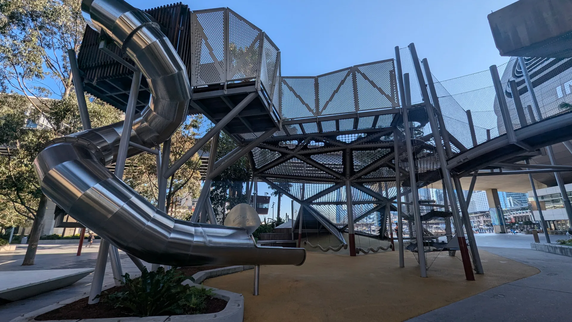 Large stainless steel spiral slide and complex rope climbing structure on a soft rubber ground, located in a shaded area with industrial architecture.