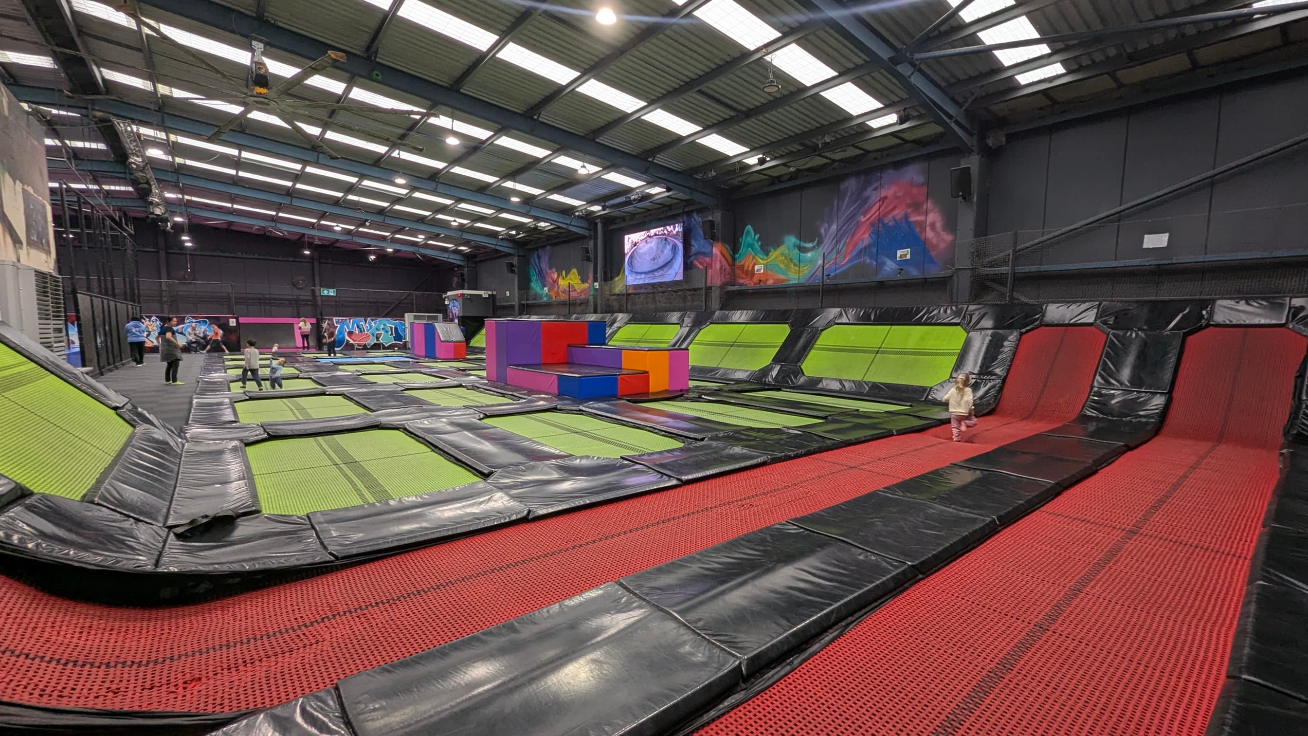 Large colourful area in a warehouse like building, with many interconnected trampolines, soft climbing structures to jump off. 