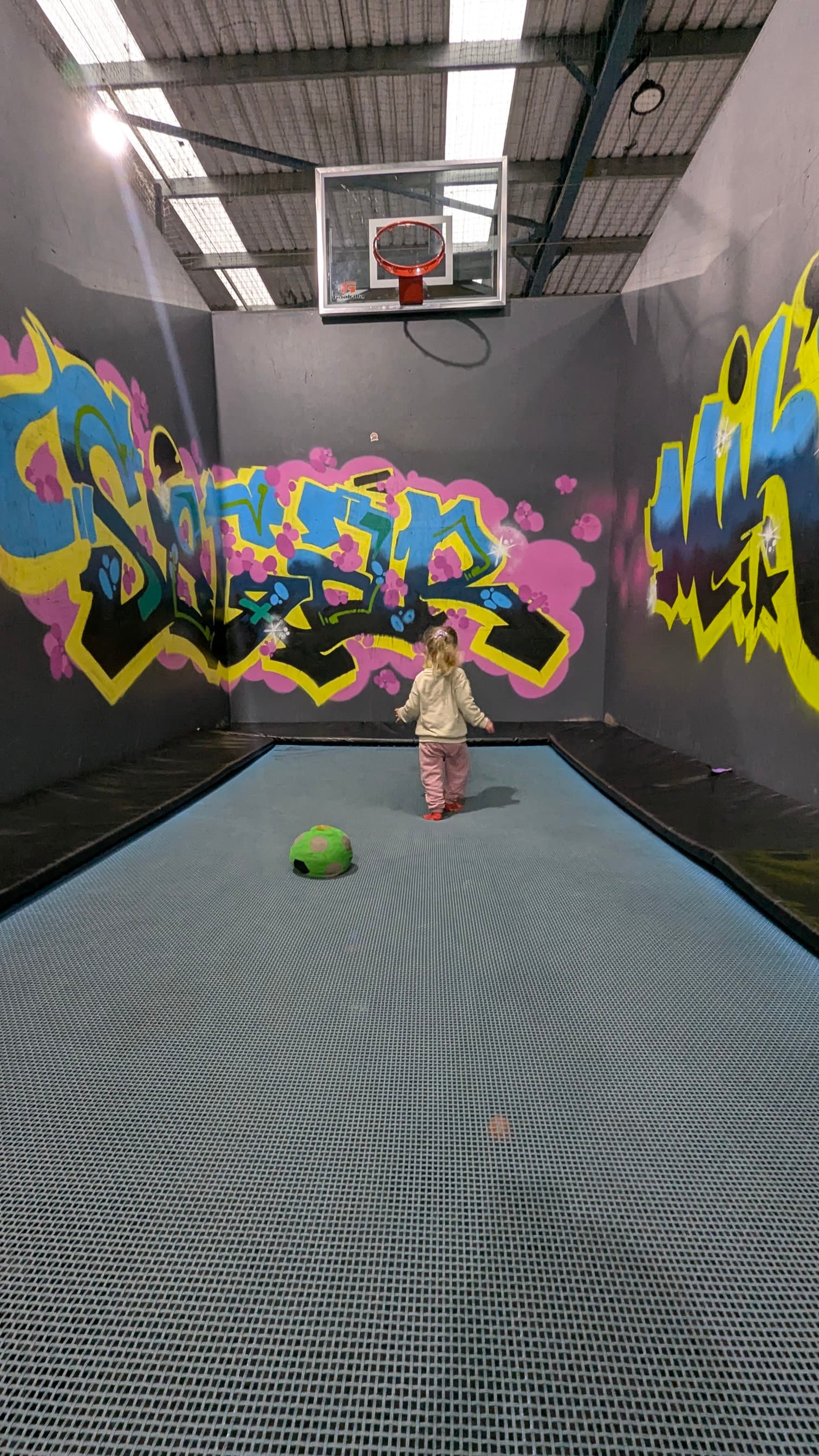 Small single trampoline surrounded by walls with graffiti like artwork and a basketball hoop.