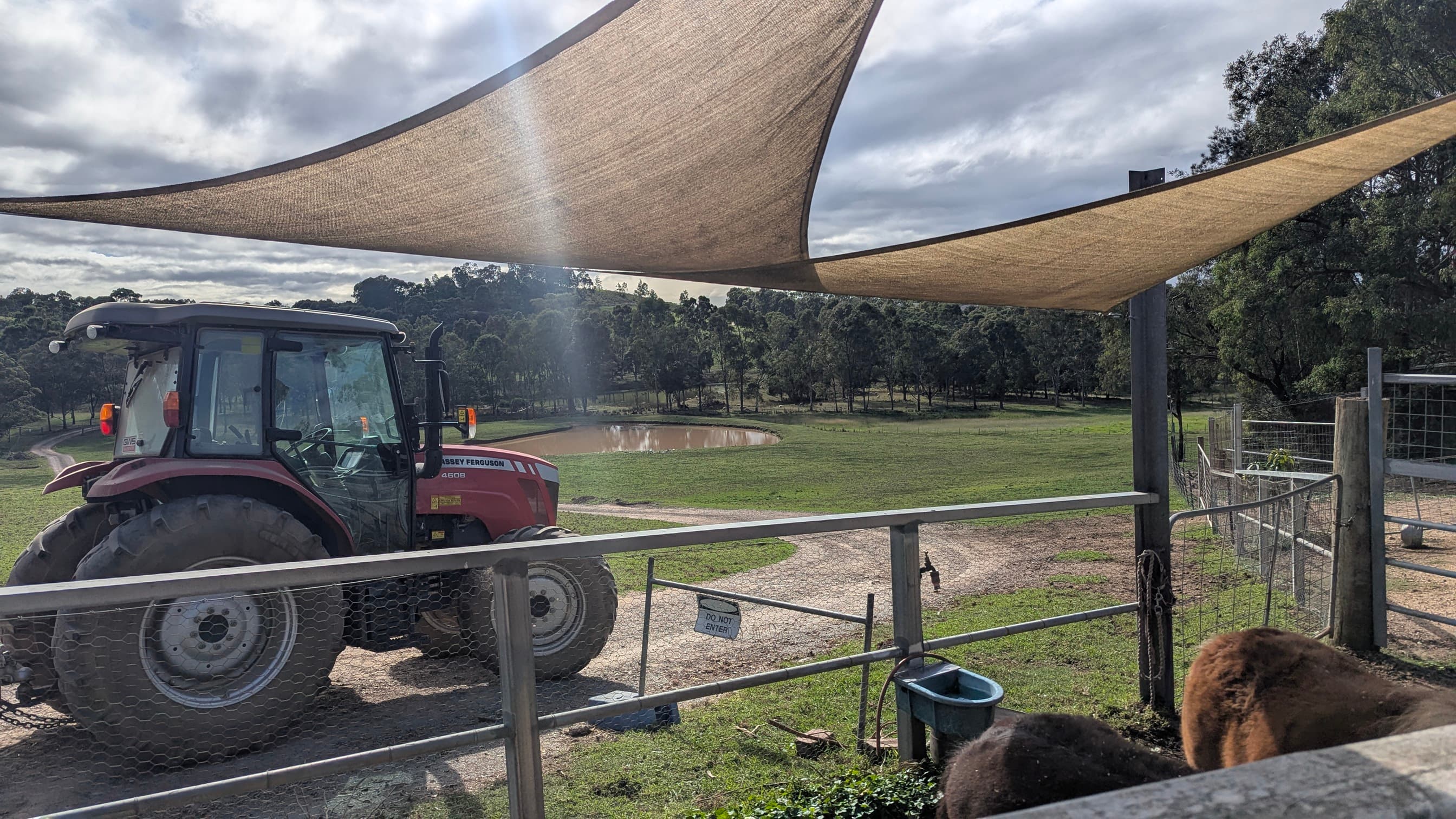 Red tractor parked under a shade sail with a field and pond in the background.