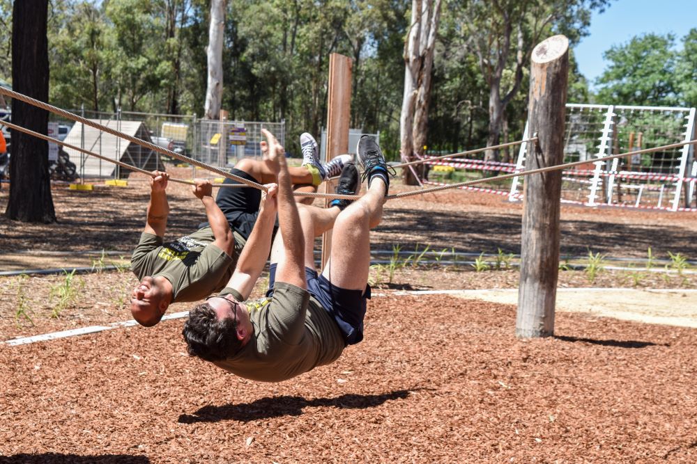 Two adults navigate a low-to-the-ground rope traverse over a soft bark mulch surface in a shaded outdoor fitness area.