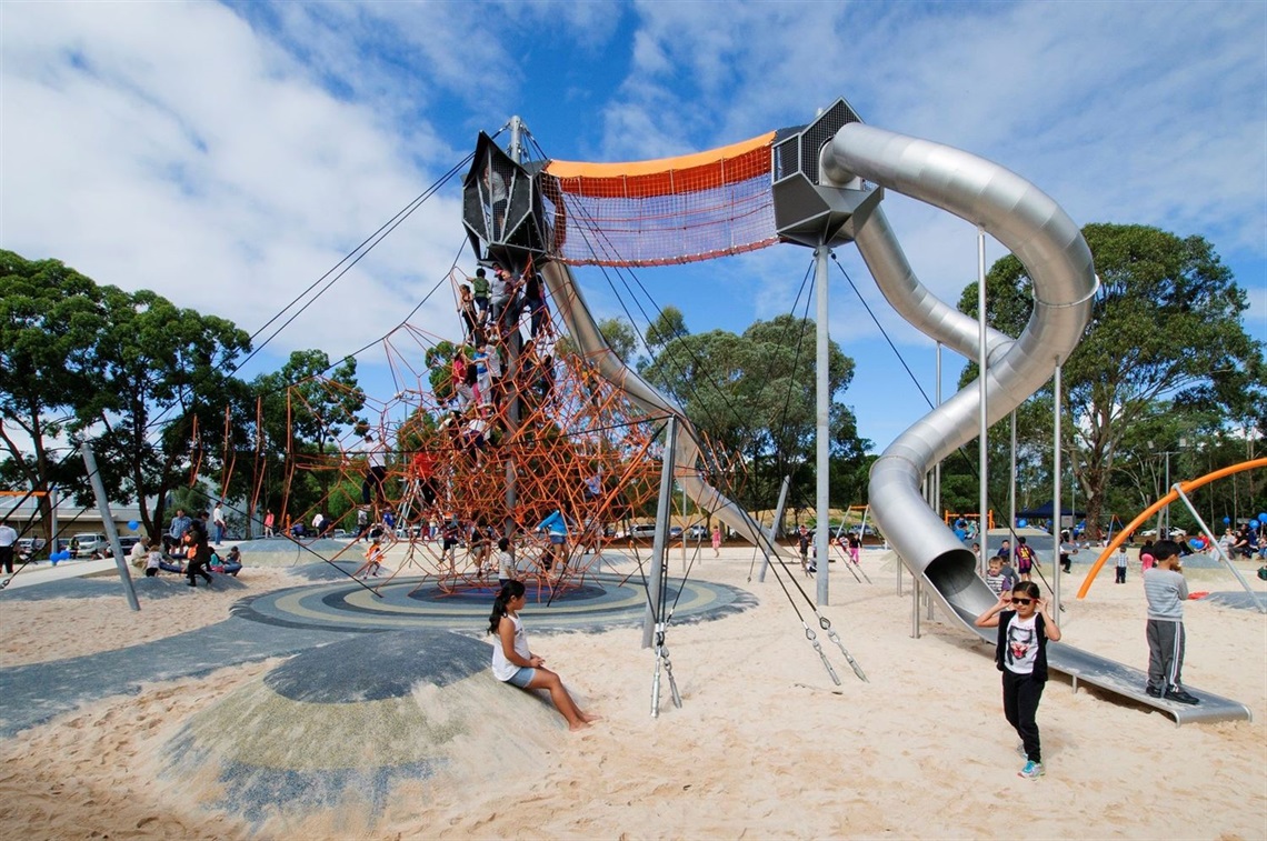 Children climb a large orange rope pyramid and use a tall, winding silver tube slide in a sand-based playground under a blue sky.