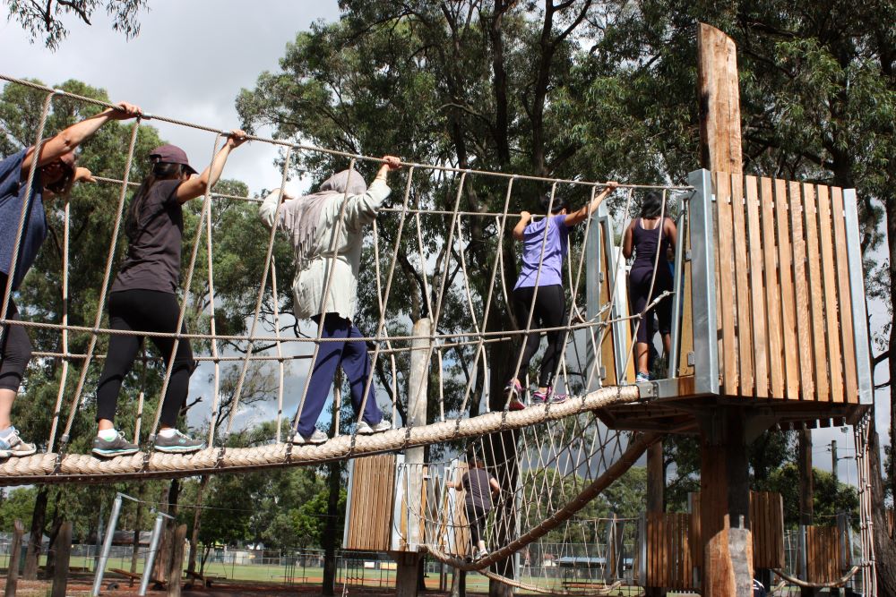 People cross a high-level rope bridge with wooden handrails and safety netting at an outdoor adventure course surrounded by tall trees.