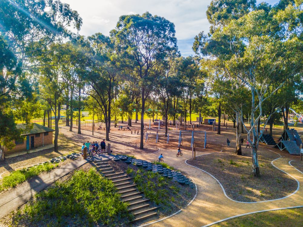 Aerial view of a sun-drenched wooded park featuring paved stroller-friendly paths, a bicycle pump track, and various timber obstacle course elements.