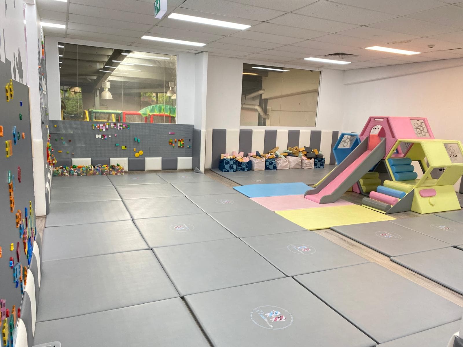 Soft play area with grey padded mats, a pink and yellow slide, and a Lego wall