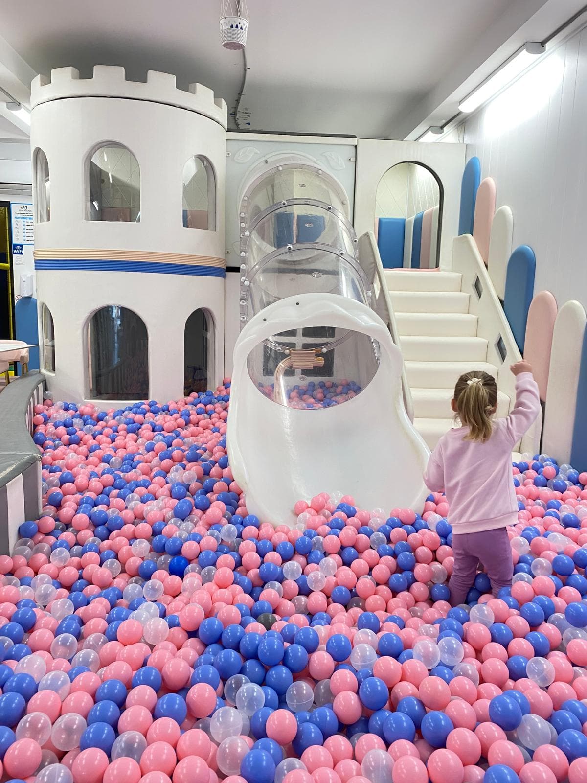 Child standing in a ball pit with pink, blue, and clear balls, next to a white play castle with a clear slide.