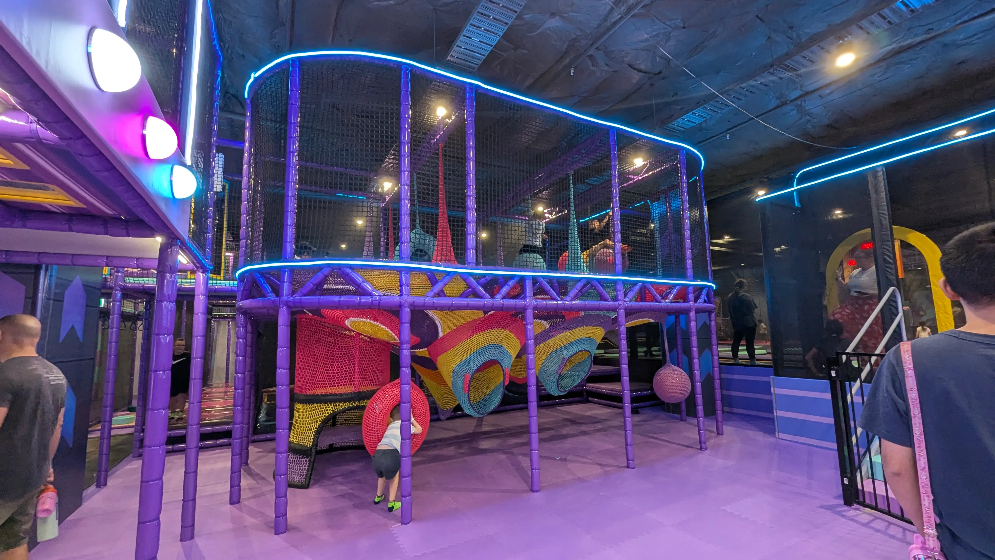 Vibrant purple multi-level play structure with colorful netted tunnels, hanging pods, and soft rubber mat flooring.