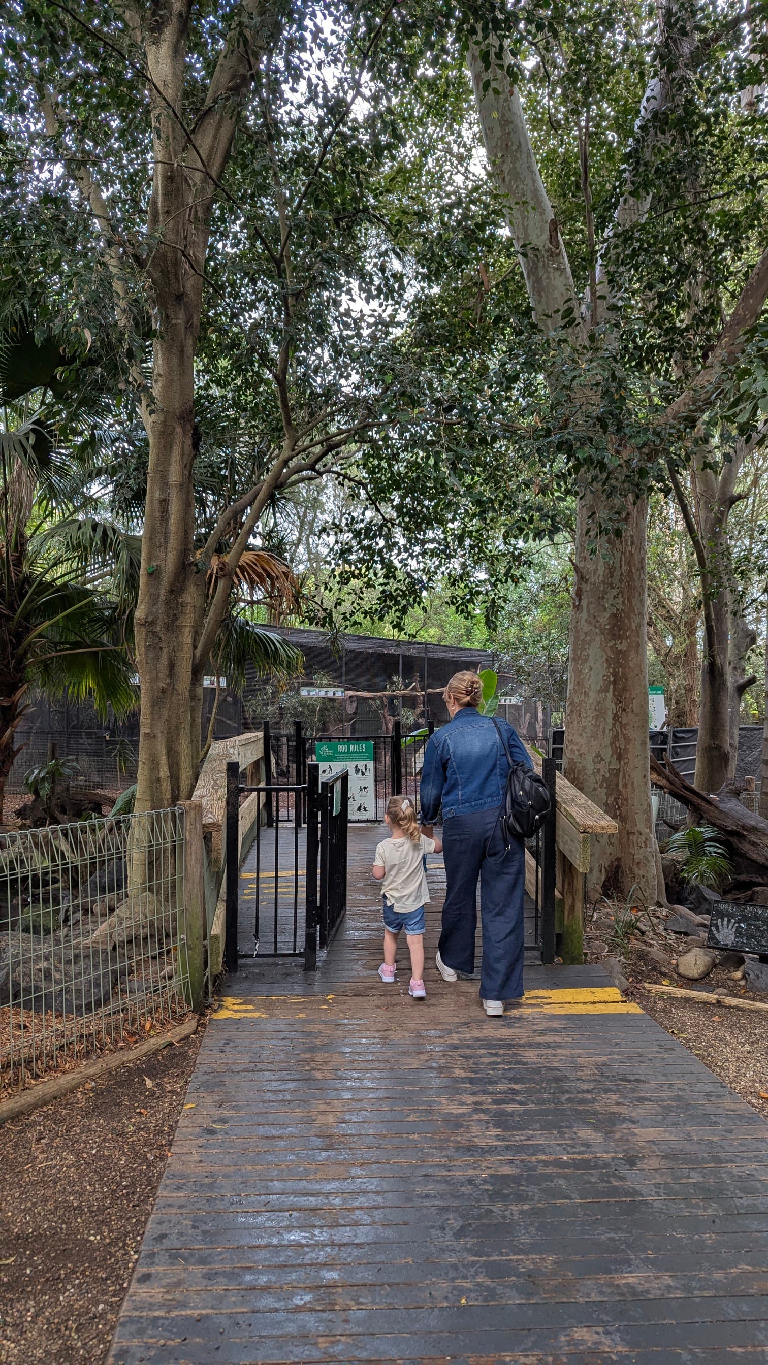 A woman and a young child walking towards the entrance of the "Wallaby Walkabout" area on a wooden bridge, surrounded by trees.