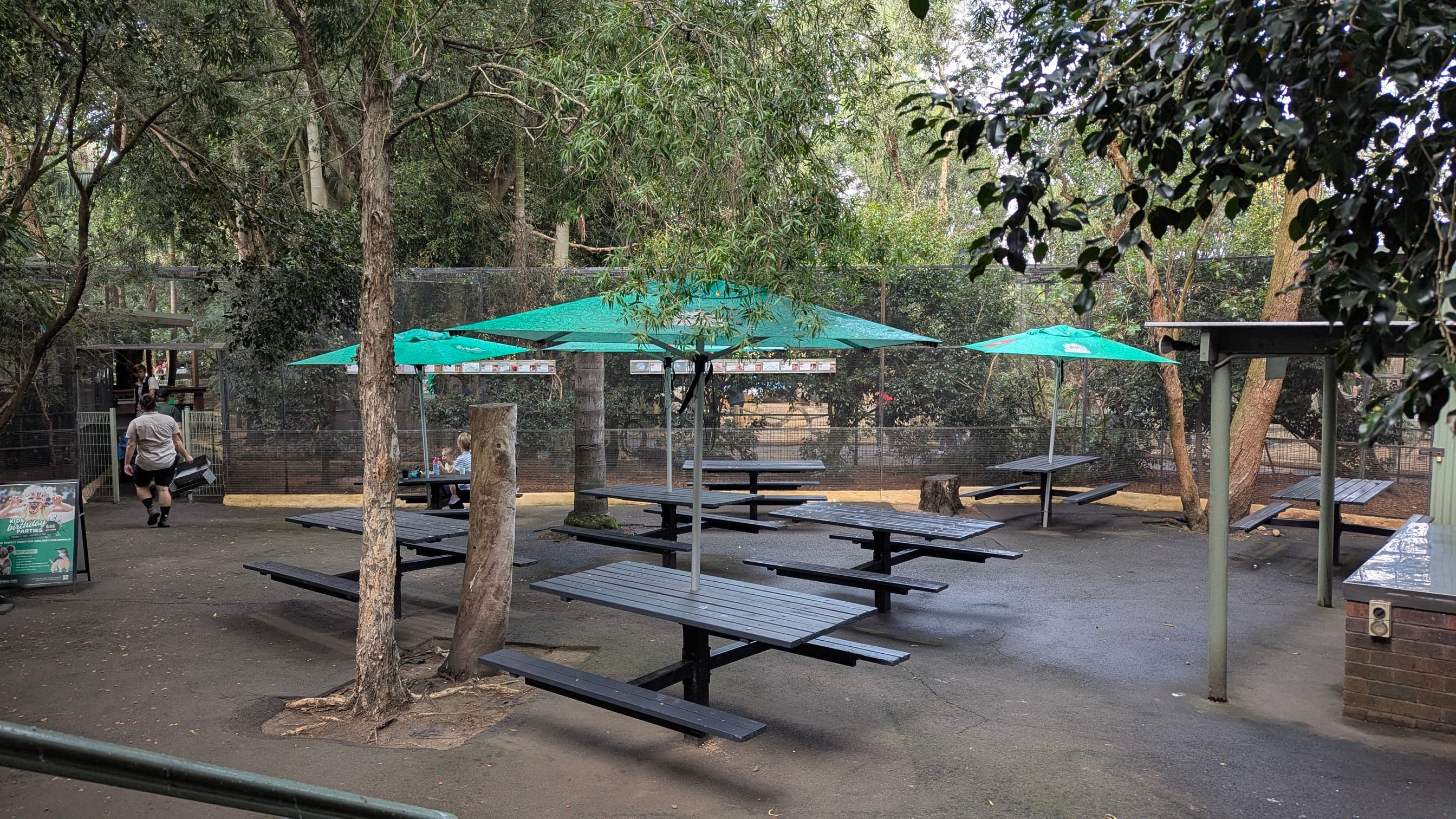 Outdoor picnic area with multiple wooden tables and benches, shaded by green umbrellas and trees.