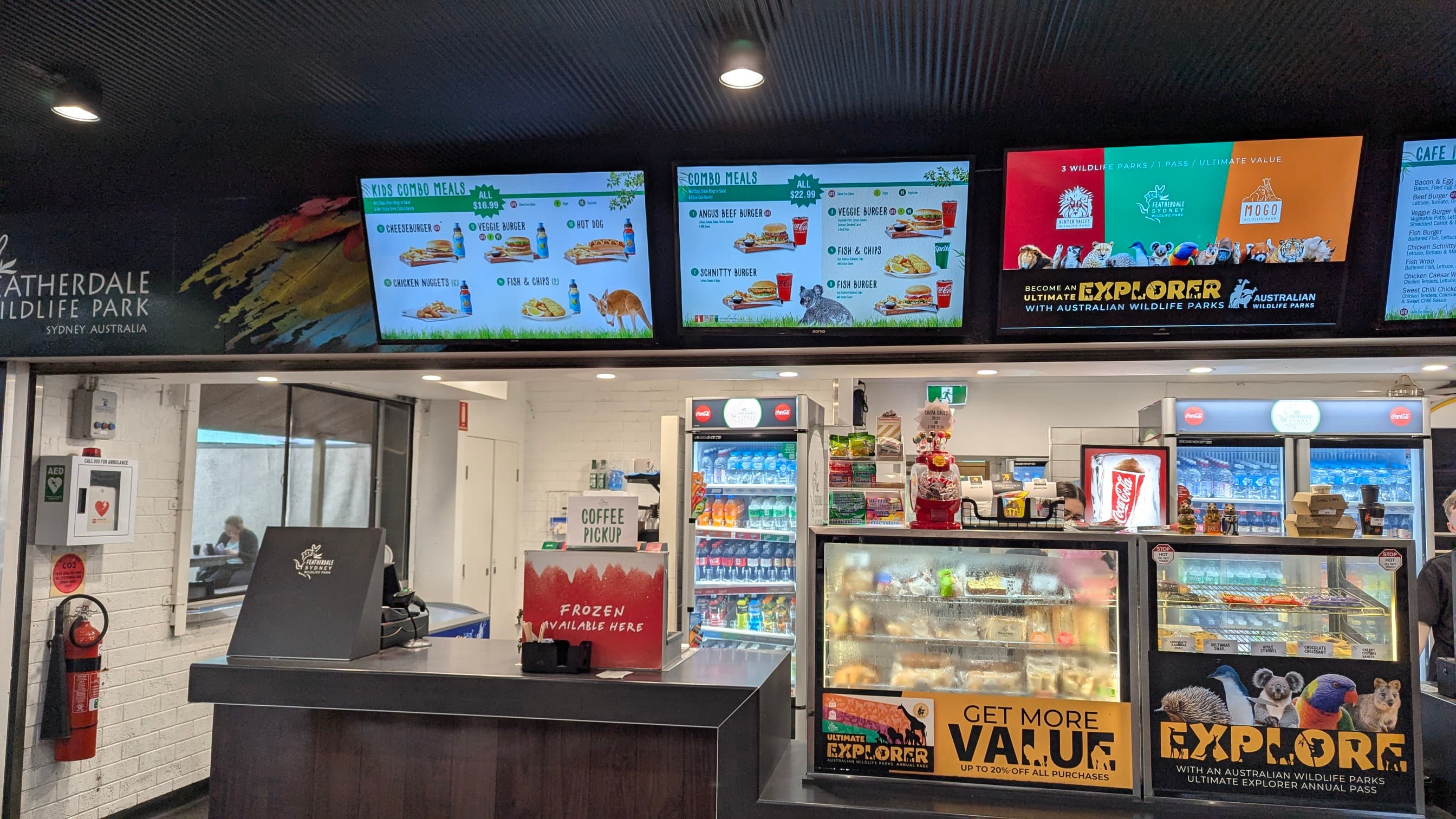 Interior of a cafe with digital menu boards showing food items, a counter, and refrigerated display cases with drinks and snacks.