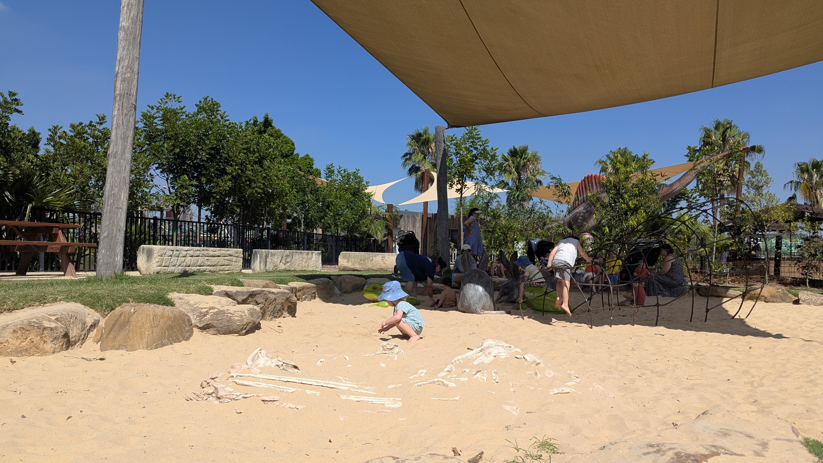Sand pit playground with a shade cloth and a small metal climbing structure.
