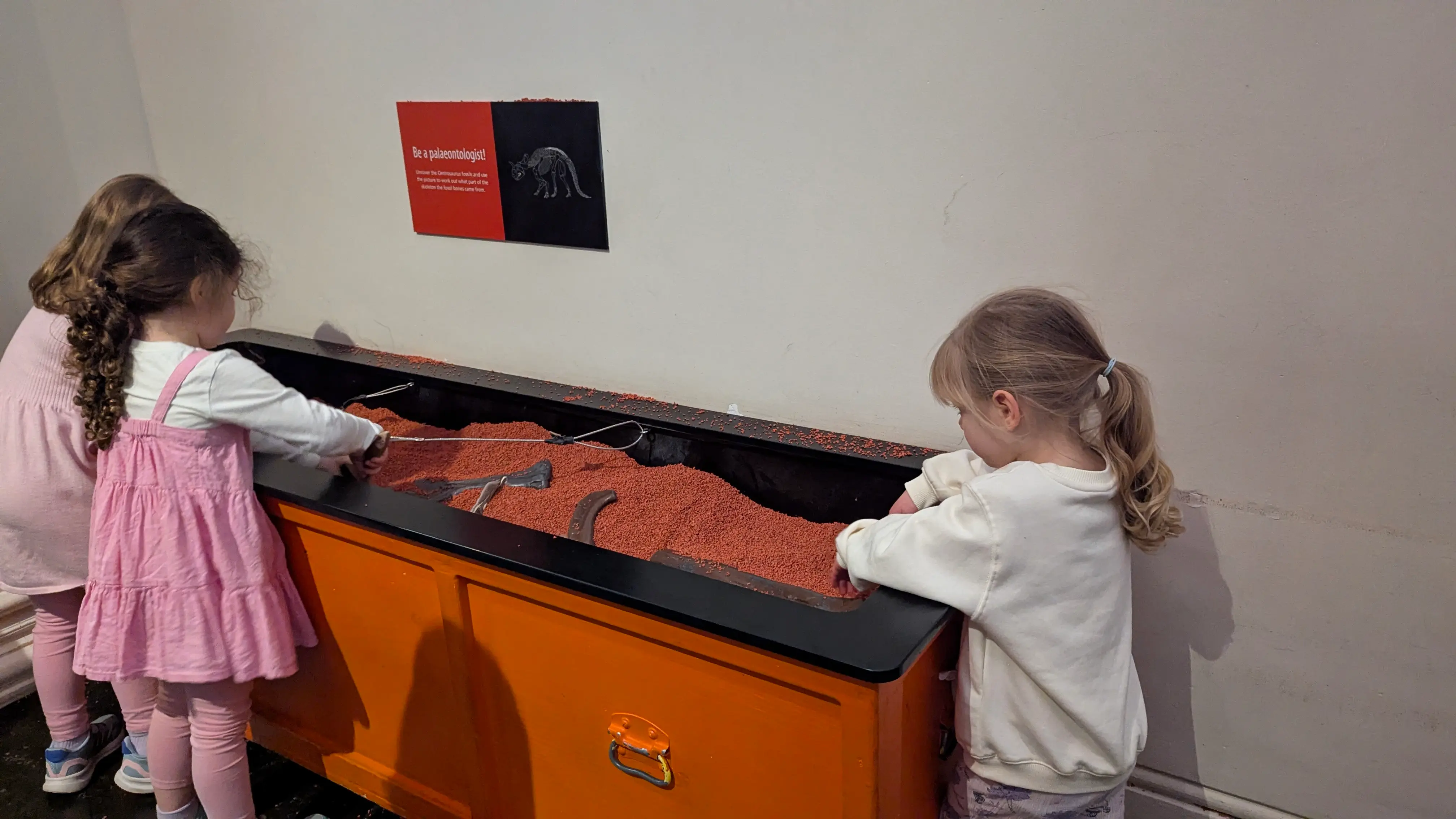 Two young children using a "Be a Palaeontologist" sand pit to dig for fossil replicas in a museum exhibit.