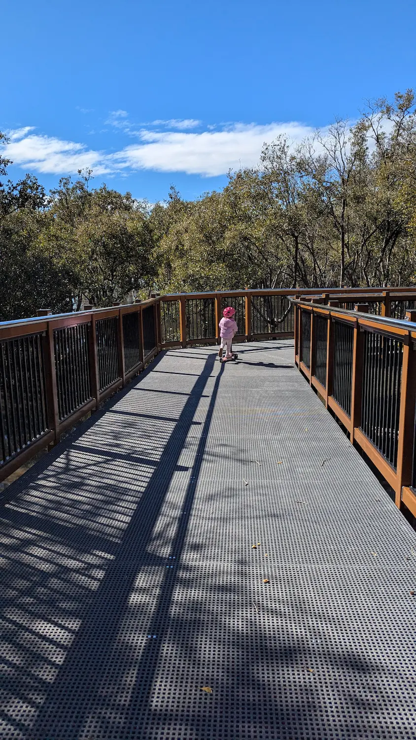 A child in a pink helmet riding a scooter on a wide, metal-grated boardwalk with safety railings, surrounded by lush green trees.