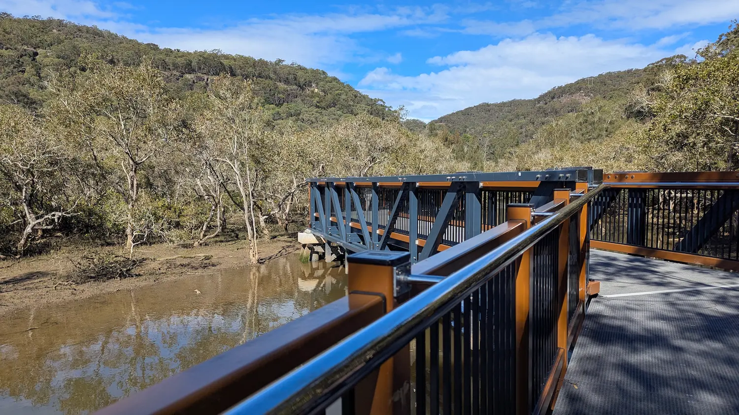 A modern pedestrian bridge section of the boardwalk passing over a muddy estuary with mangrove trees under a blue sky.