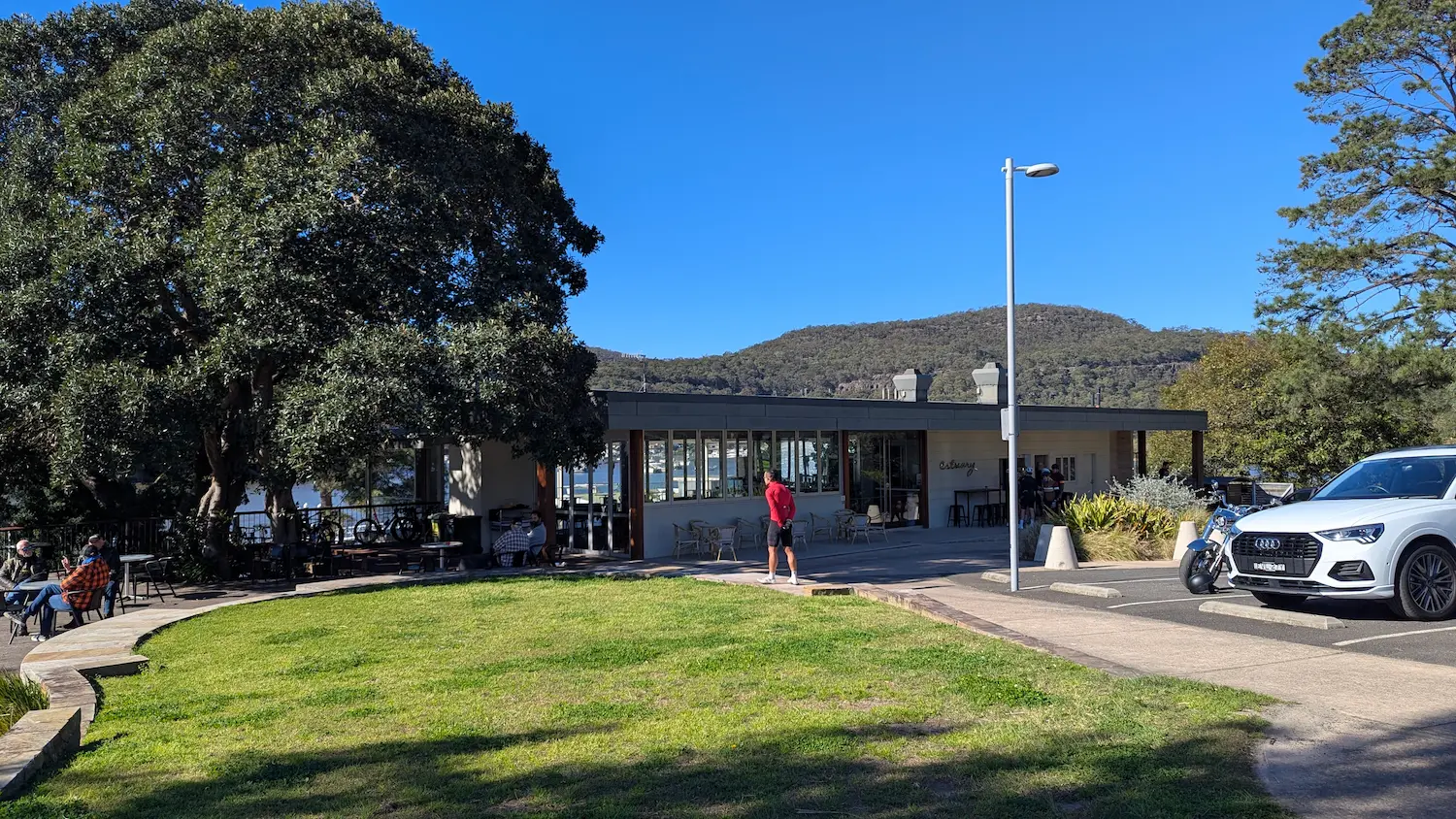 Estuary Cafe with outdoor seating under a large shade tree, featuring flat paved access and a nearby grassy area.