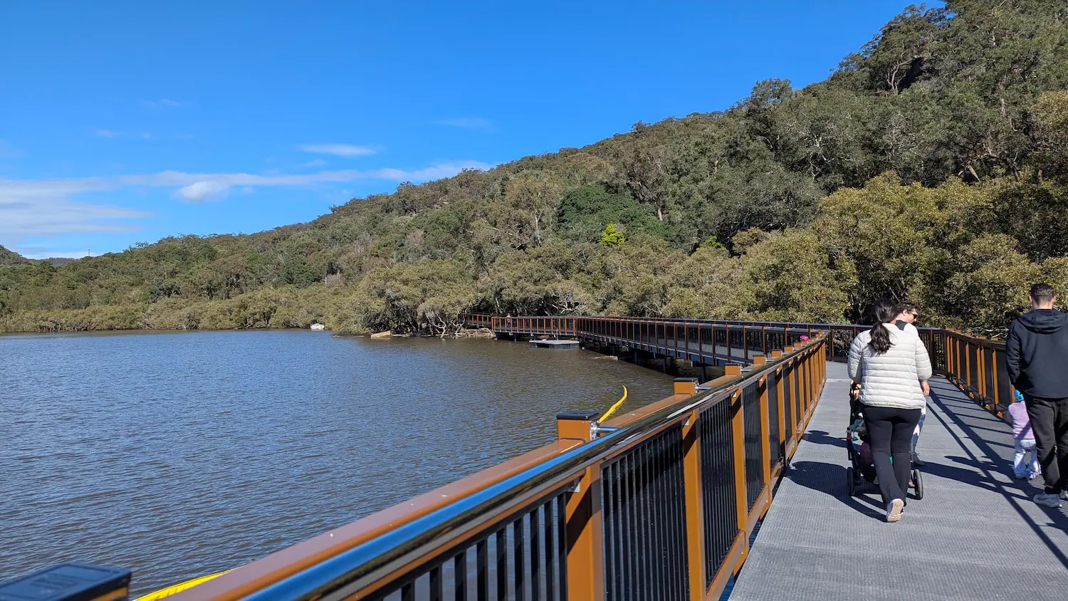 A stroller-friendly, accessible boardwalk curving along the water's edge, featuring high safety railings and views of a forested hillside.