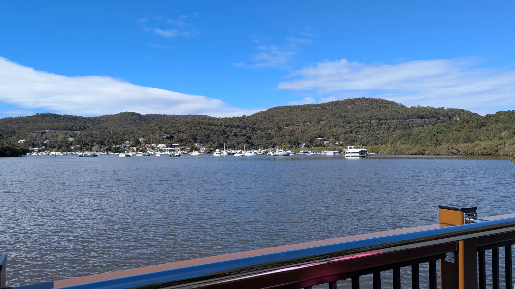 Scenic view from the boardwalk overlooking a wide river filled with white boats and yachts, backed by rolling green hills.