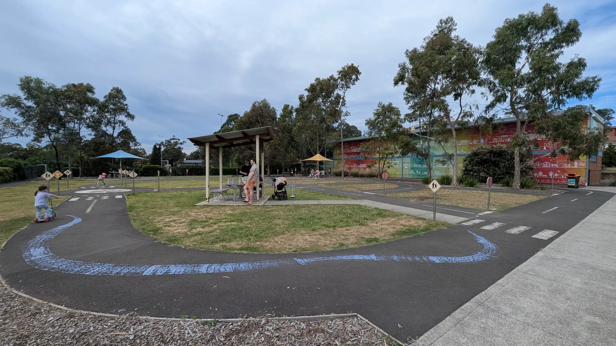 A paved kids' scooter track with road markings, a sheltered picnic table, and a colorful mural wall under mature trees.