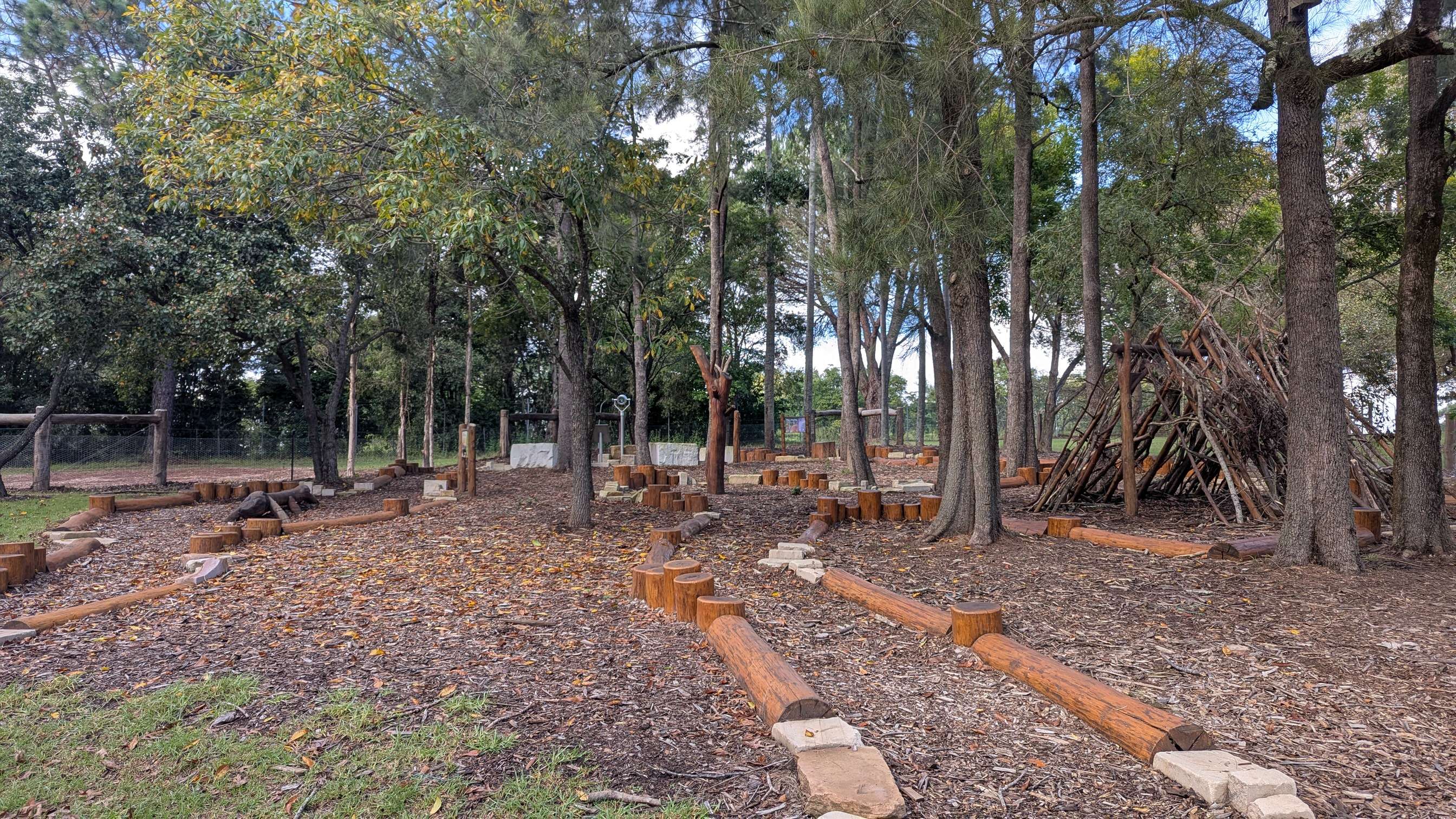 Forest themed playground set amongst tall trees with a stick hut, lots of wooden logs and stumps to balance on.