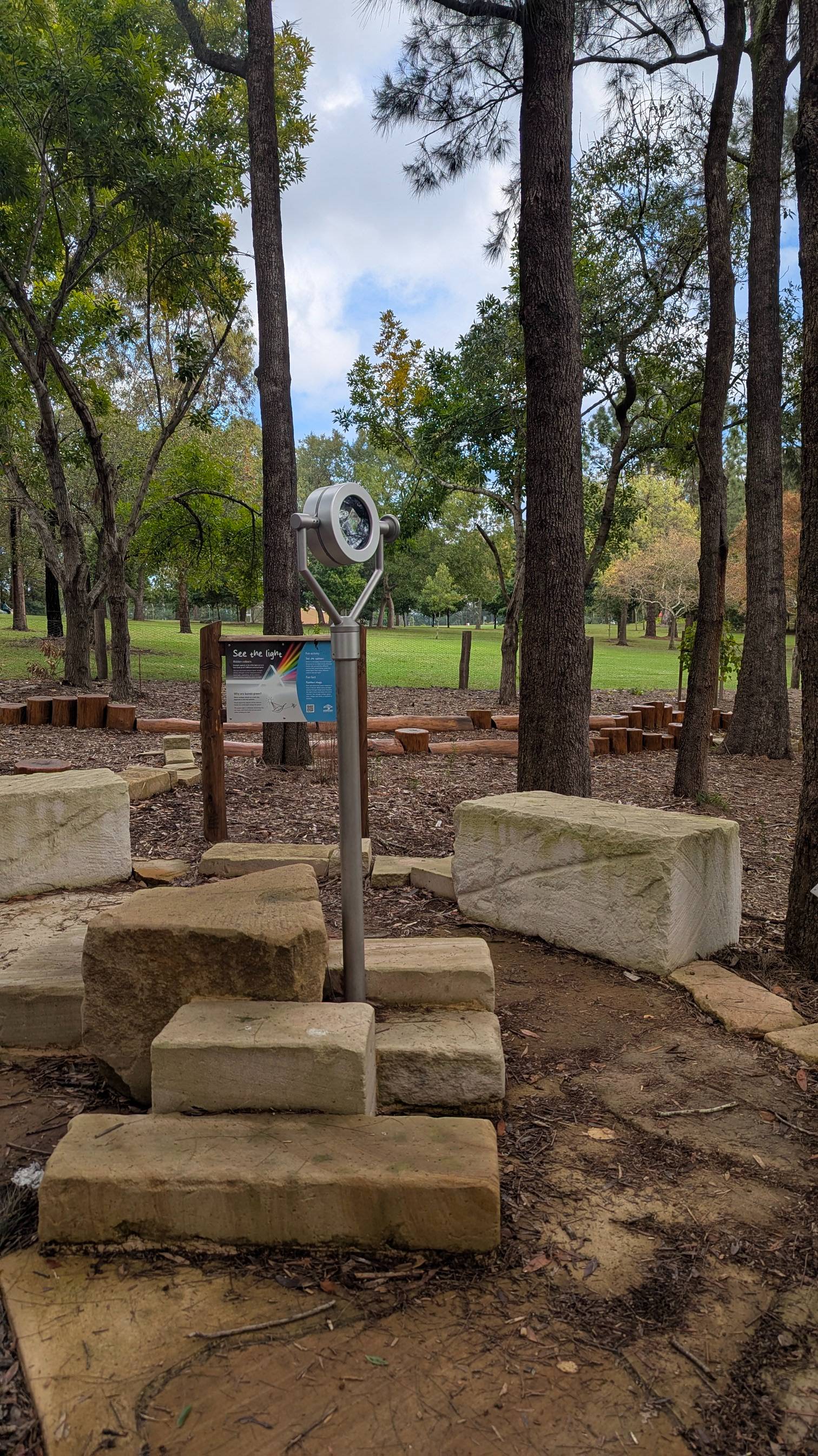 Alt text: Outdoor prism exhibit on a metal stand with a “See the light” sign, surrounded by stone blocks and trees.