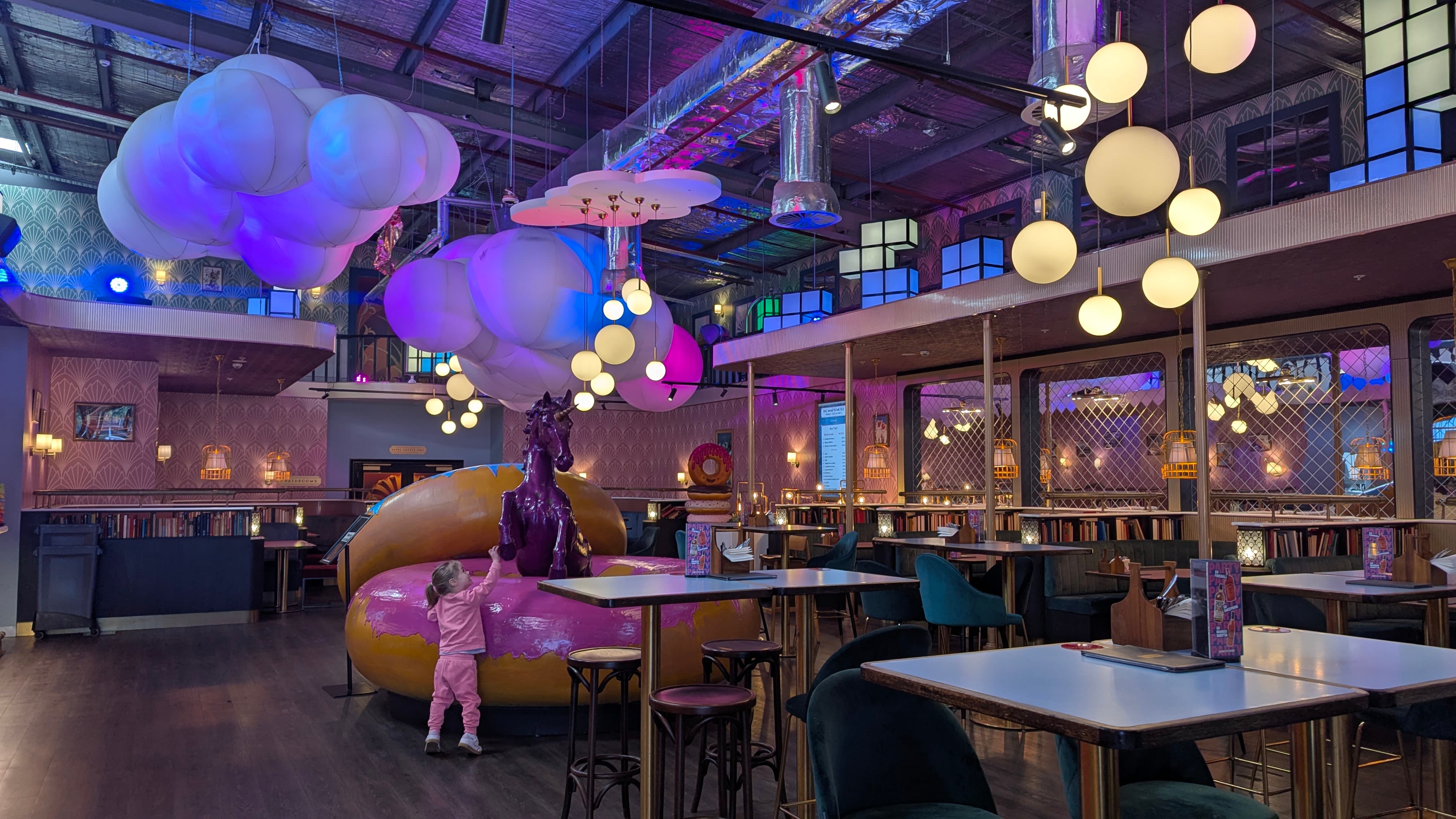 A vibrant, modern lobby interior with a child reaching towards a large donut sculpture, plush cloud decorations, and various seating areas.