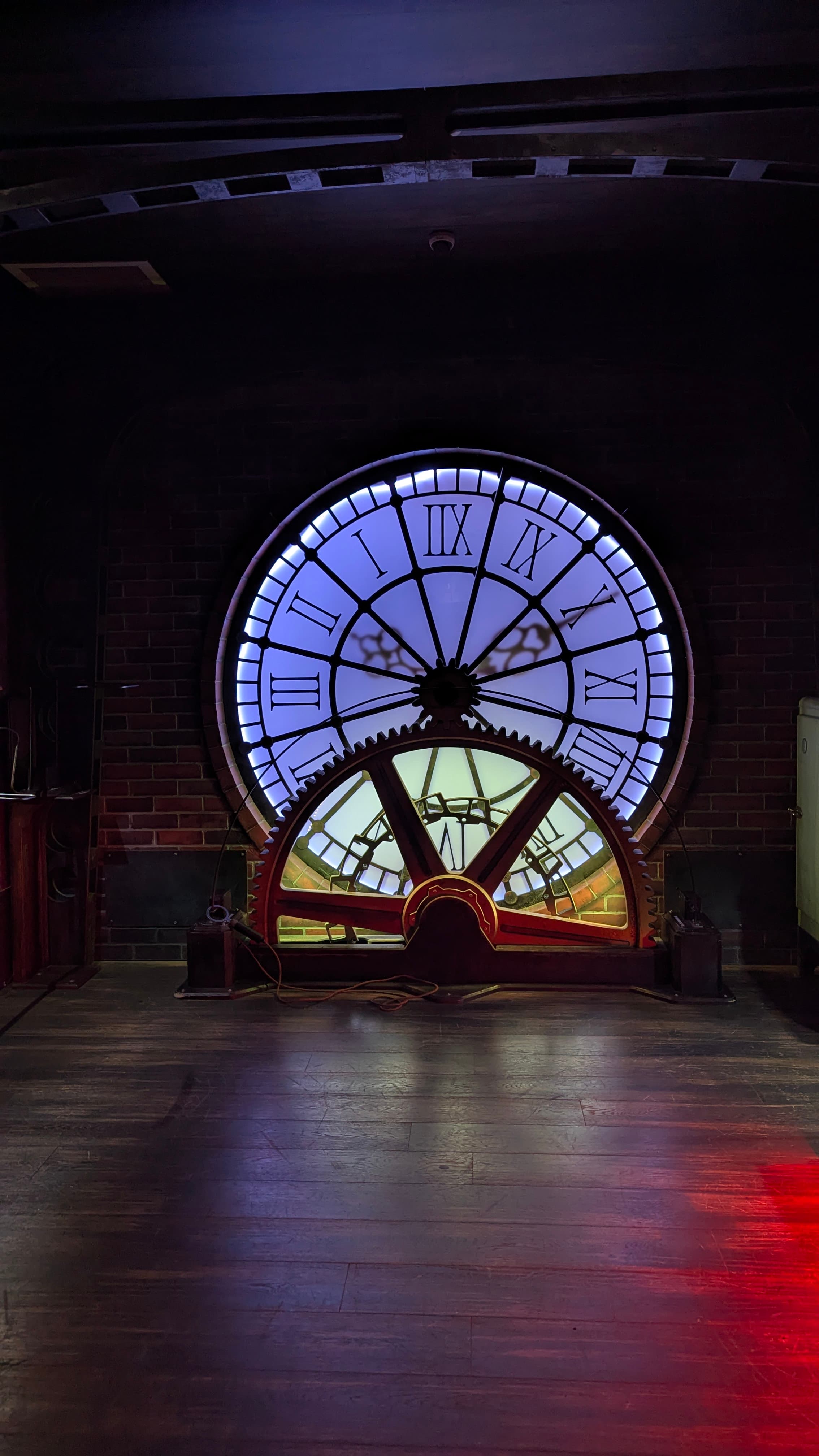 A large, illuminated clock face with Roman numerals and a prominent red gear in a dimly lit room, suggesting a steampunk or industrial theme.
