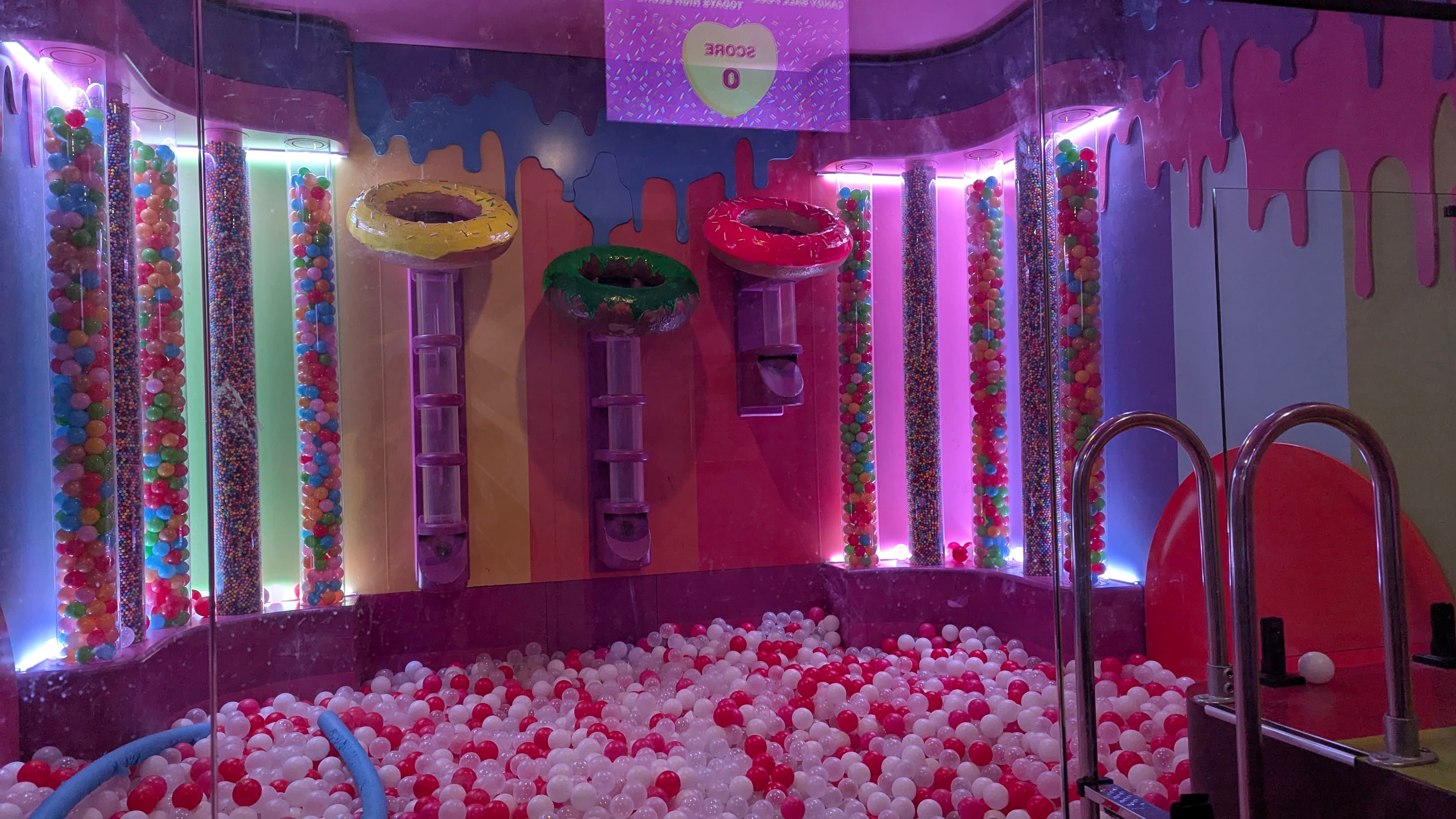 A colorful indoor ball pit filled with white and red balls, featuring giant sweet-themed decorations and bright lights.