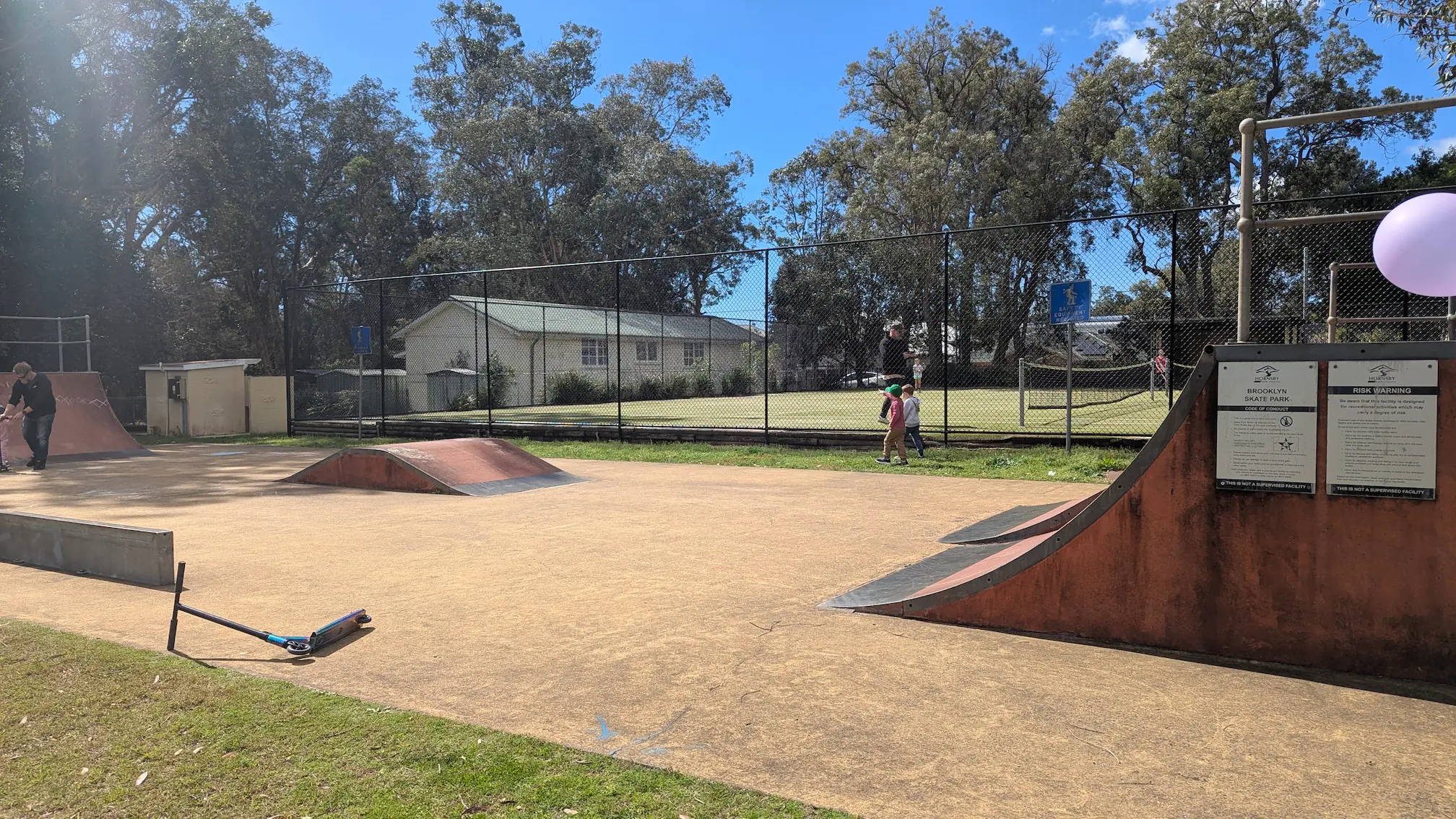 Brooklyn Skate Park featuring a concrete flat ground, small ramps, and a quarter pipe. A scooter lies in the foreground near safety signage.