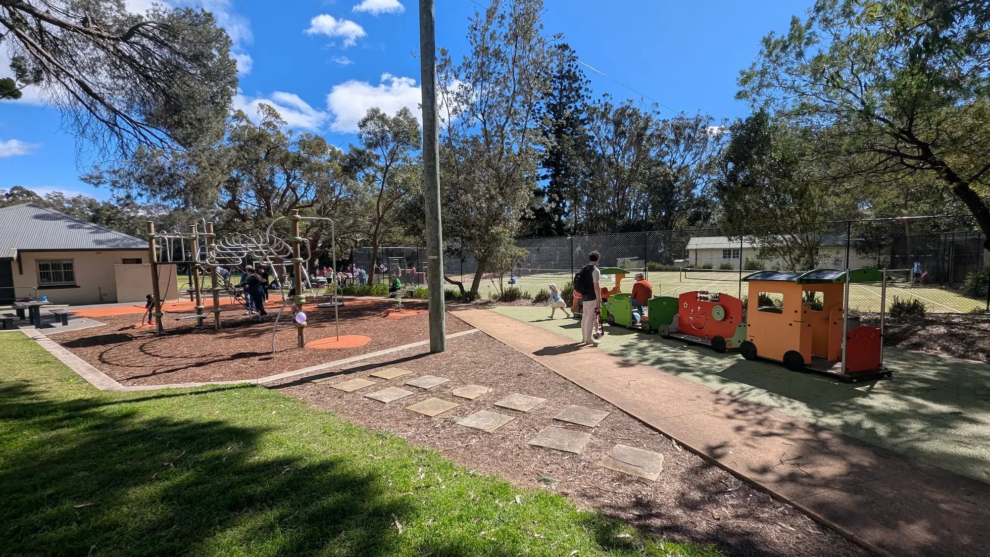 Sun-drenched playground with a climbing frame on mulch and a play train on rubber flooring, featuring paved paths for stroller access.