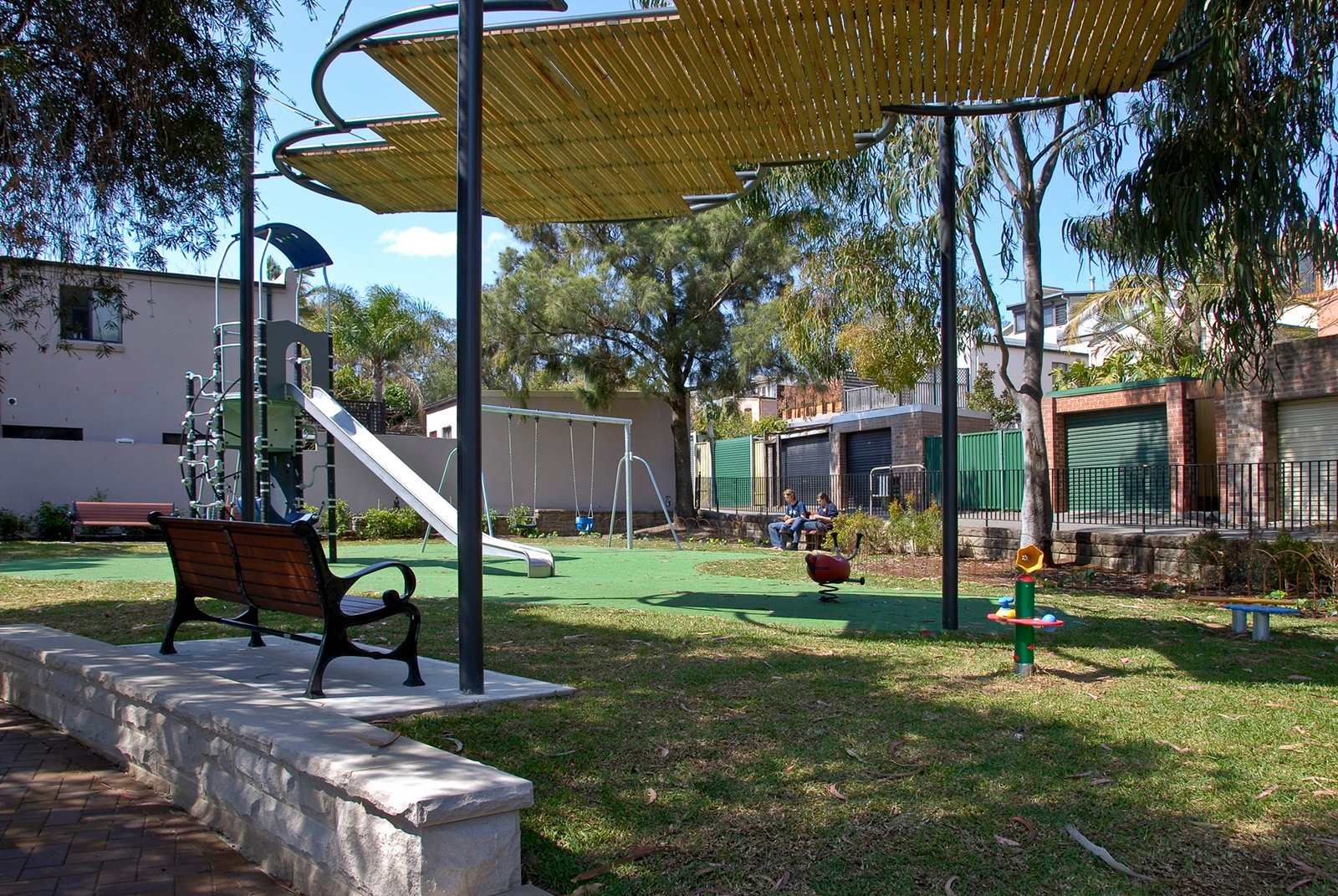 Shaded playground at Charles Kernan Reserve with a bench, swings, and other play equipment.