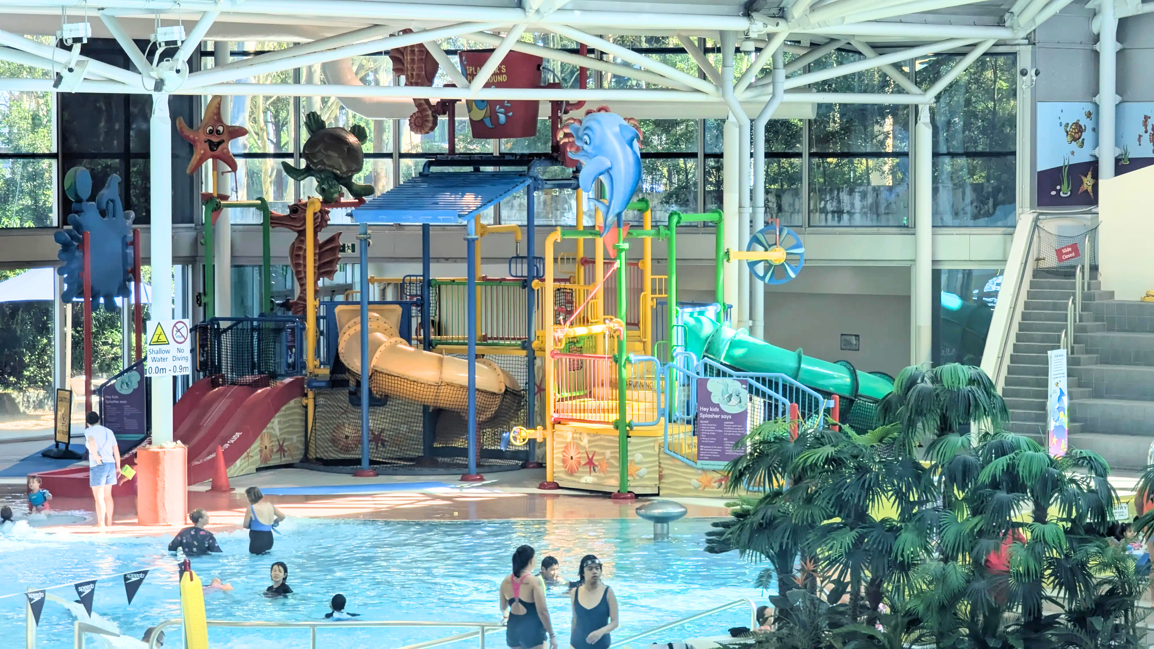 "Splasher’s Playground" featuring a gated entry, multiple small slides, climbing structures, and a giant tipping bucket. Includes a rubber-matted splash zone and shallow entry pool.
