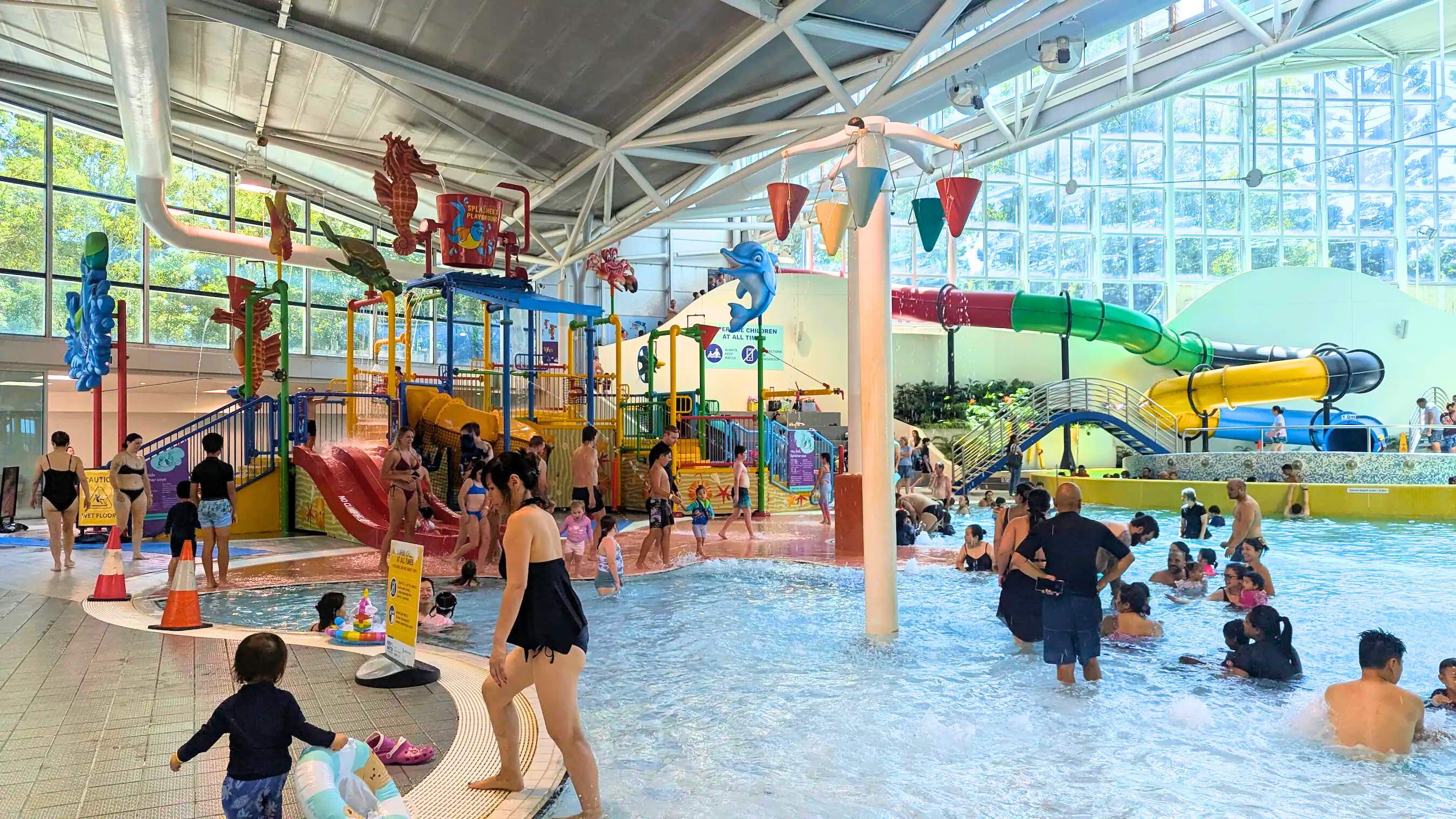 Vibrant indoor splash pad with shallow water, bucket drenchers, sea-themed slides, and various tipping buckets. Families are wading in the pool under a bright, high-ceilinged roof.