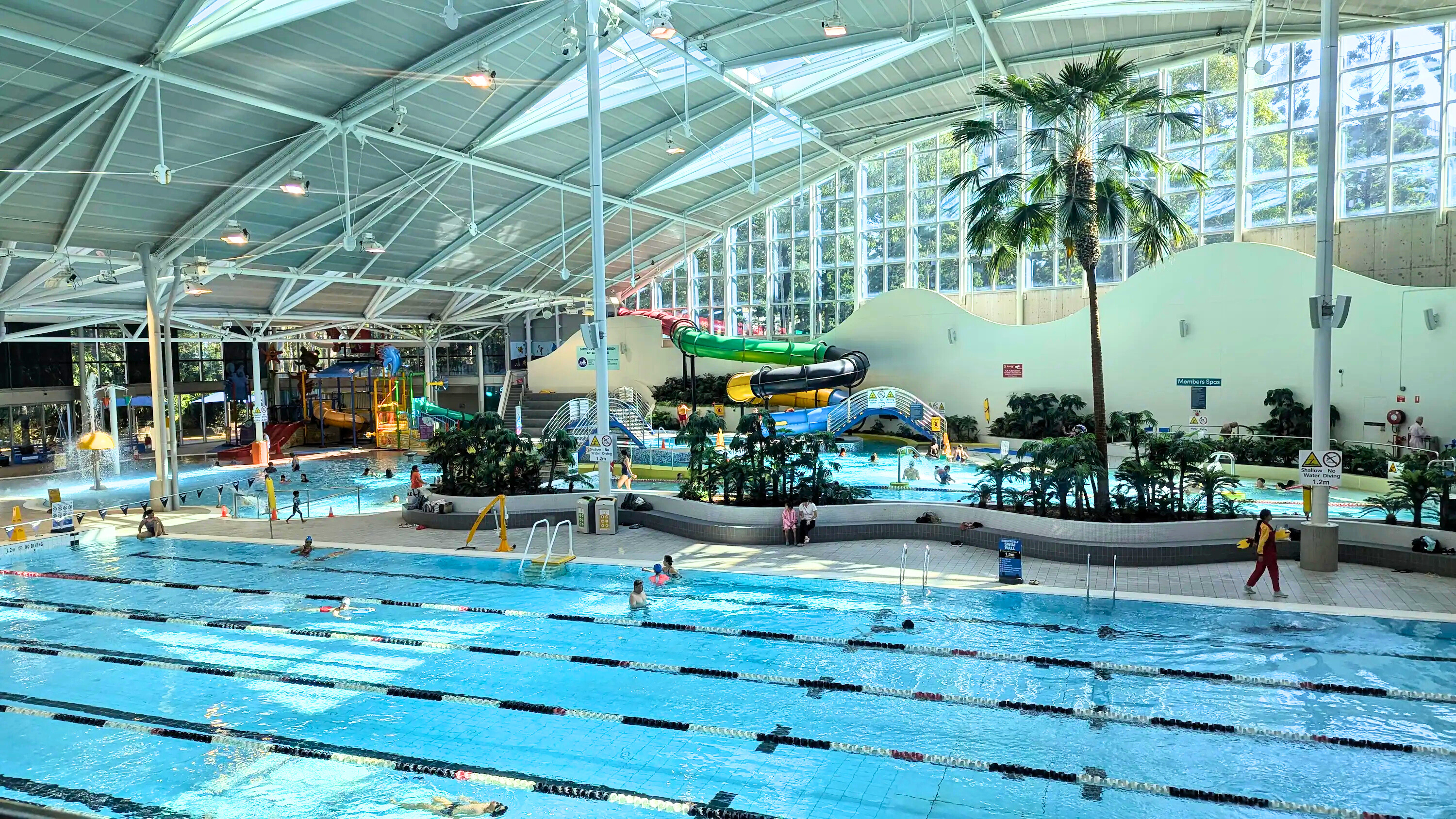 Wide view of an indoor aquatic center showing lap lanes, a tropical-themed leisure pool with palm trees, and large colorful water slides in the background.