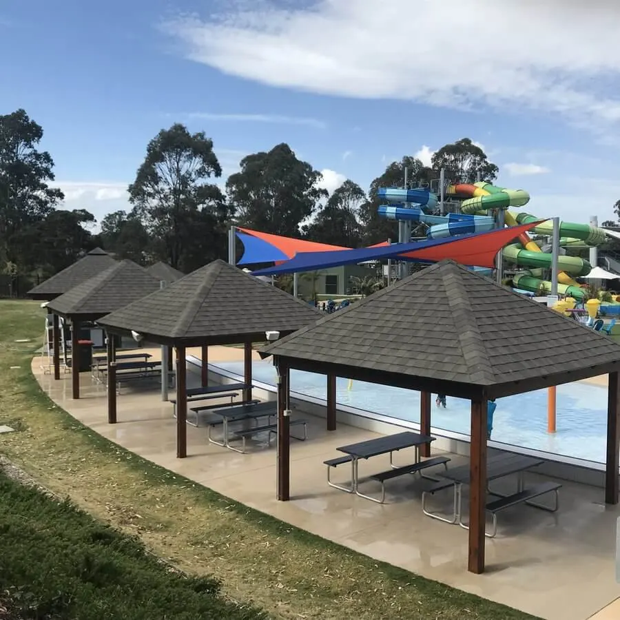 A row of wooden cabanas with dark shingled roofs and picnic tables, situated on a concrete path overlooking a splash pad and colorful shade sails.