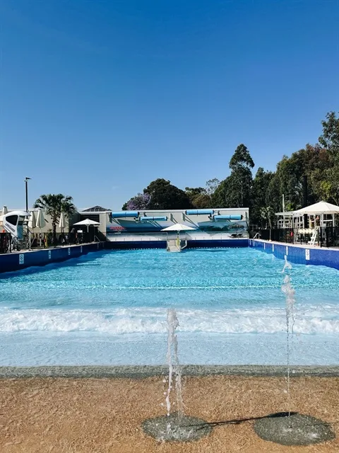 A wide, bright blue wave pool with gentle ripples, a lifeguard station on the left, and a white sun umbrella in the center under a clear blue sky.