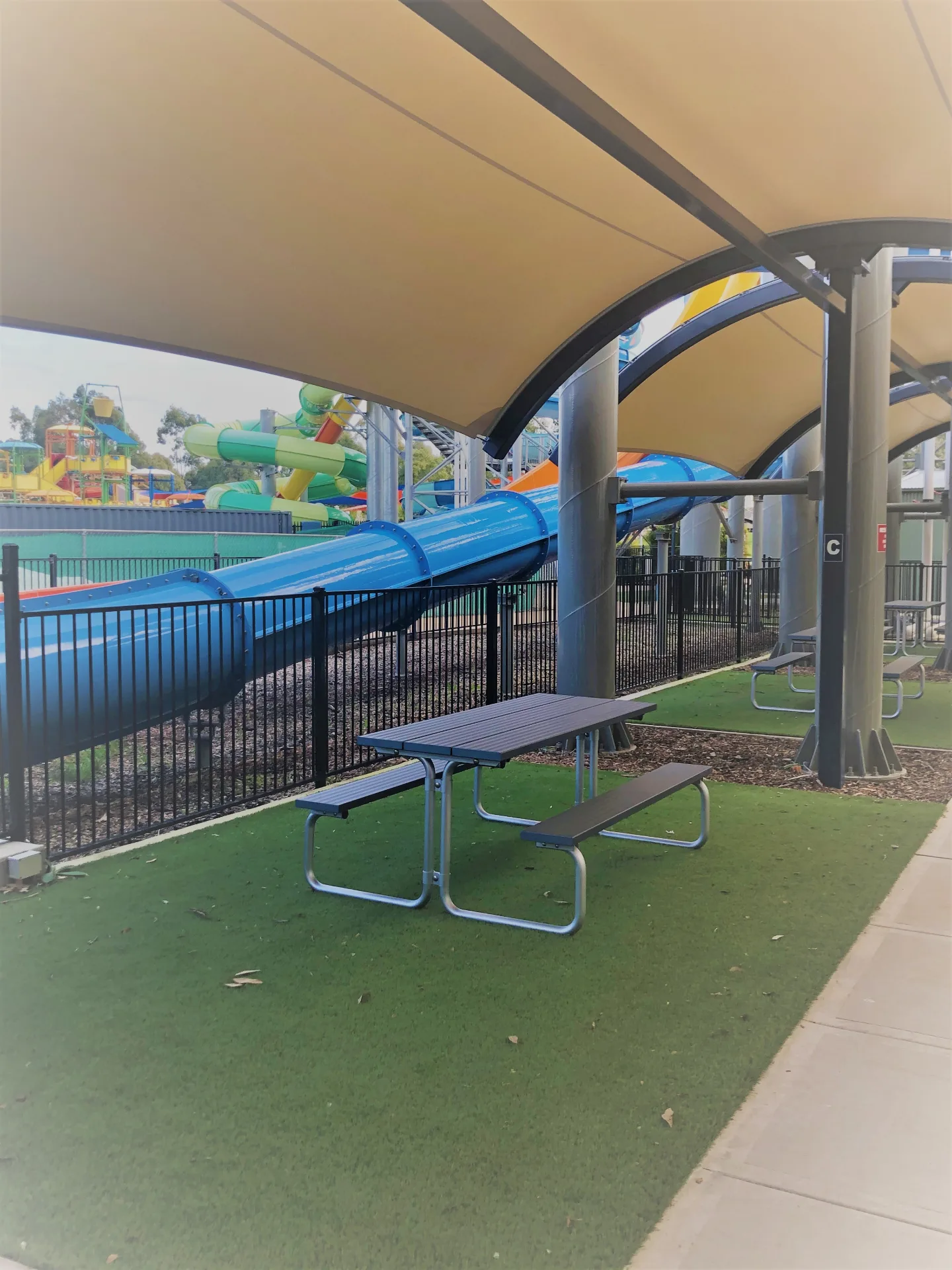 A shaded seating area with a picnic table on green artificial turf, located under a large tan canopy next to a blue water slide tube and a safety fence.