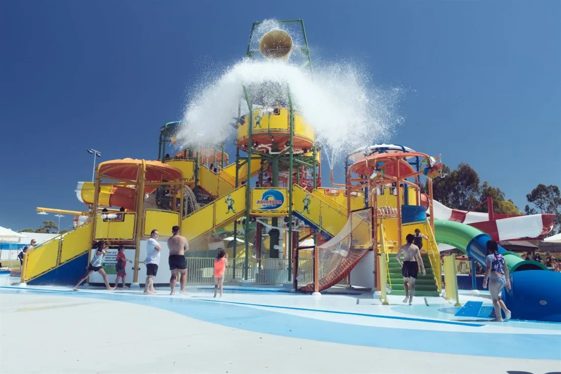 Vibrant yellow and orange multi-level water play structure with several slides and a large bucket at the top splashing water over visitors in a shallow pool.