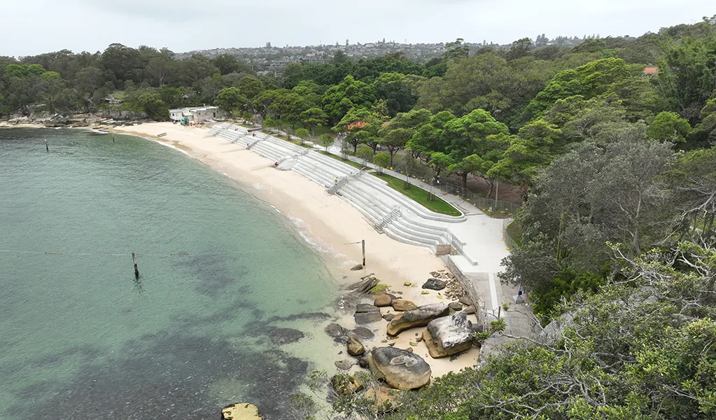 Curved sandy beach with terraced concrete steps leading to calm turquoise water, surrounded by lush green parkland and scattered rocks along the shoreline.