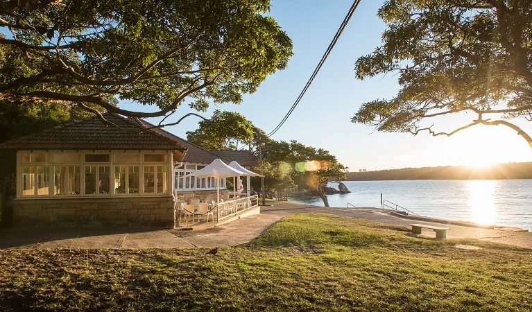 Waterfront pavilion with wraparound deck beside calm beach, surrounded by large shade trees and grassy lawn area perfect for family picnics.