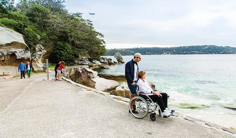 Man pushing woman in wheelchair along waterfront path at Nielsen Park, with calm harbor waters, rocky shoreline, and lush parkland visible