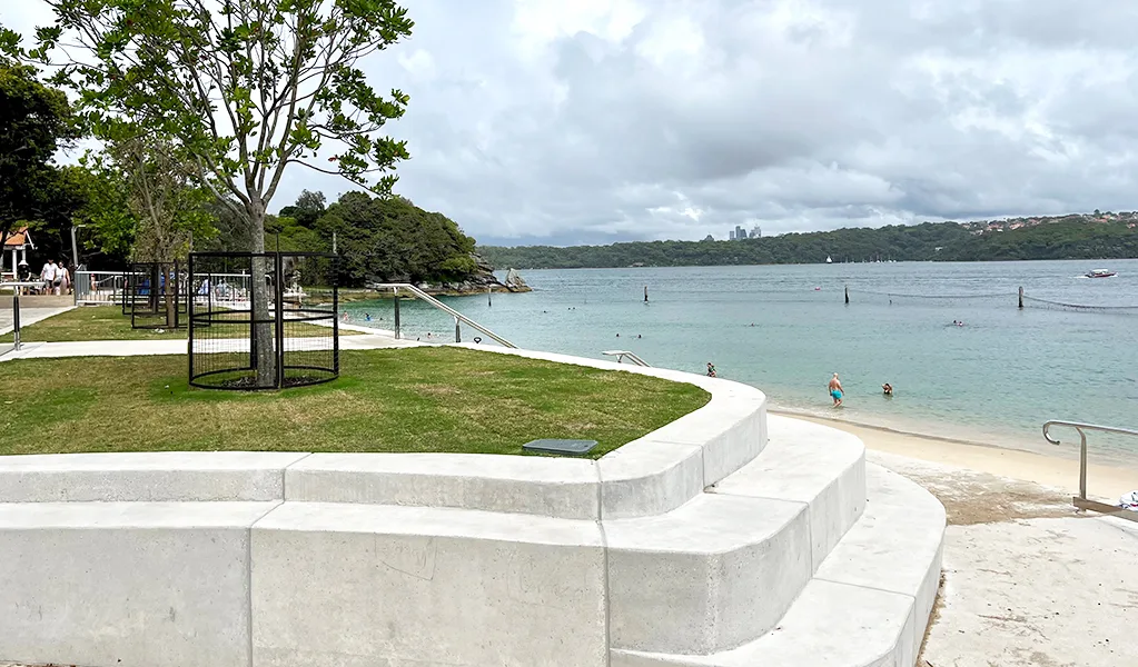Concrete terraced seating overlooks a calm beach with clear turquoise water, featuring a fenced playground area on grassy lawn with large shade trees nearby.