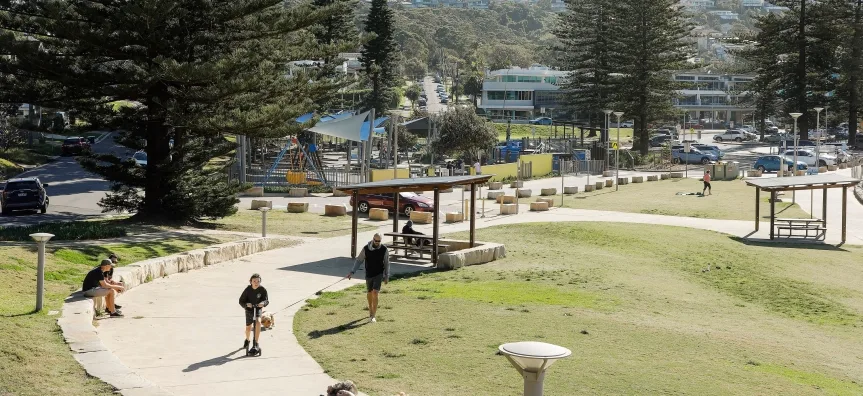 Large grassy park with covered picnic tables, paved pathways, and mature pine trees providing shade. Playground equipment visible in background with residential hillside beyond.
