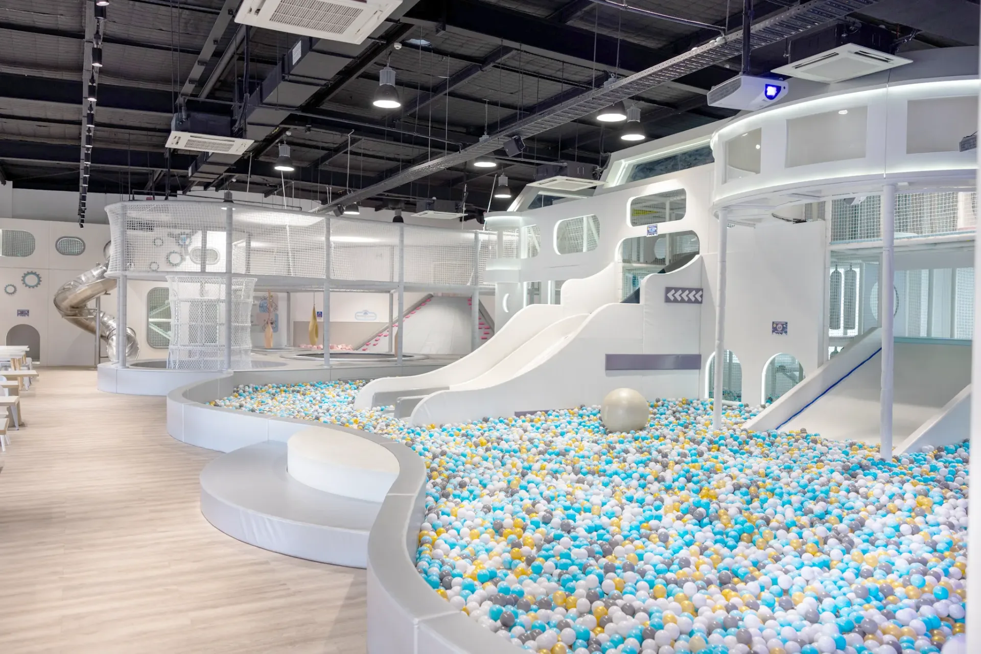 Modern white multi-level play structure with slides and climbing features, surrounded by large ball pit filled with colorful plastic balls in blue, white, yellow and gray tones in bright indoor play center.
