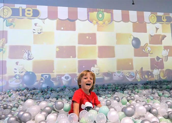 Child playing in colorful ball pit with soft play balls at indoor play center, with interactive digital wall game displaying cartoon characters in background