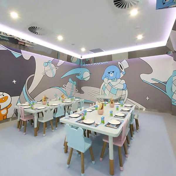 Bright children's party room with space-themed wall murals featuring cartoon astronauts and planets, white tables and colorful chairs set up for dining