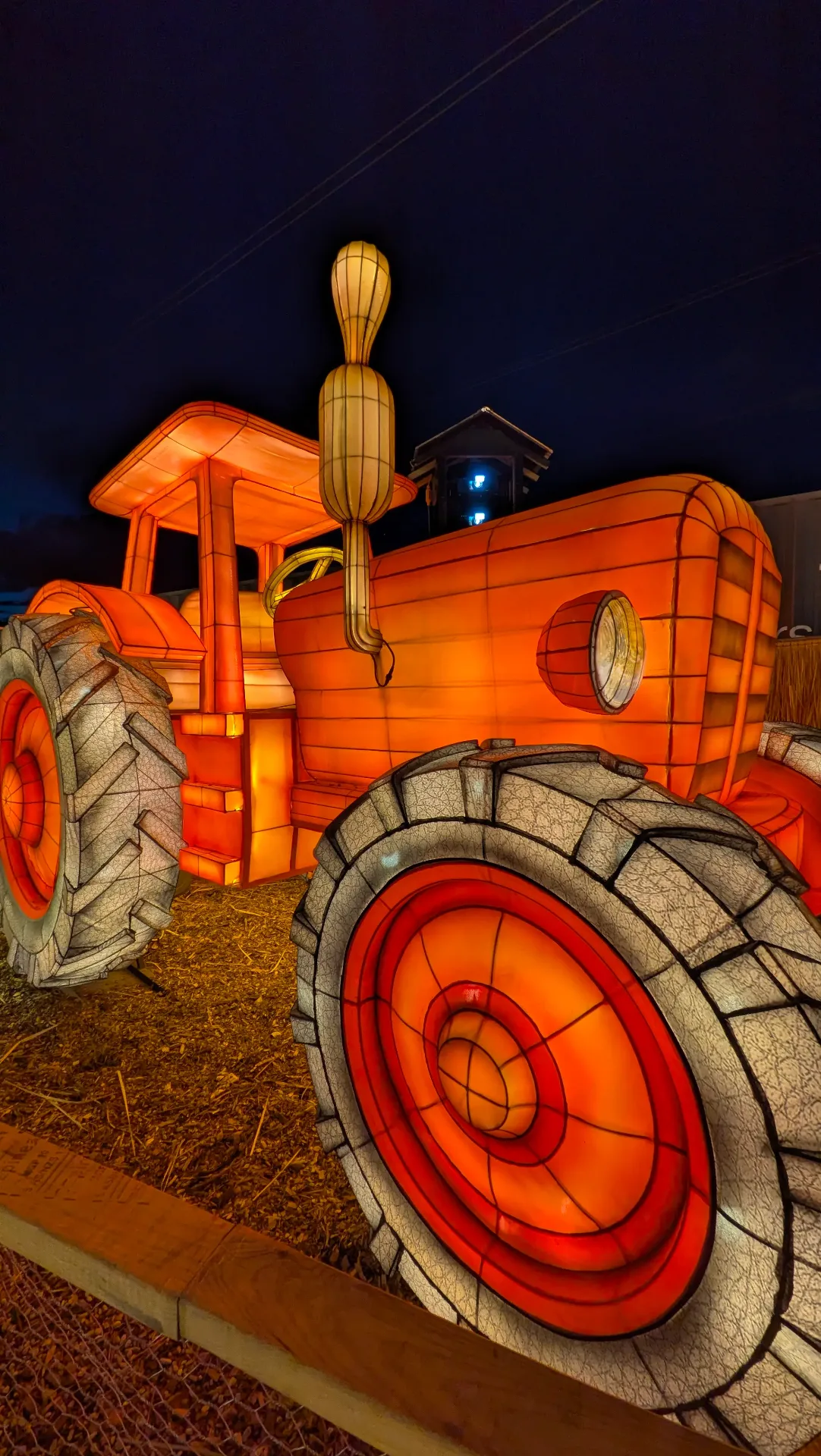 A large glowing tractor lantern sculpture in vivid orange and red, displayed on a bed of straw at the Lightfields night event. A illuminated gourd rises from the exhaust pipe against a dark evening sky.