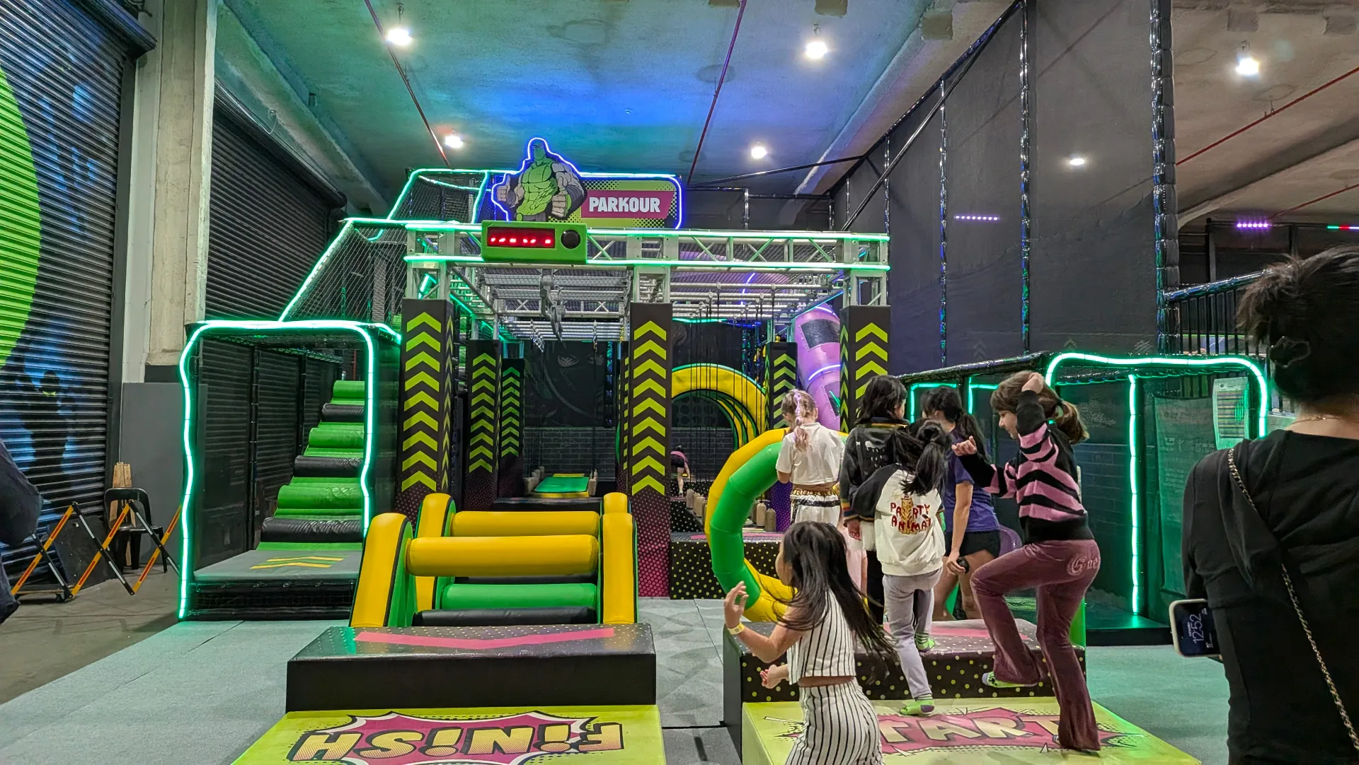 Indoor trampoline park with colorful obstacle course featuring foam blocks, padded barriers, and climbing structures. Children and adults navigate the neon-lit parkour area with safety padding throughout.