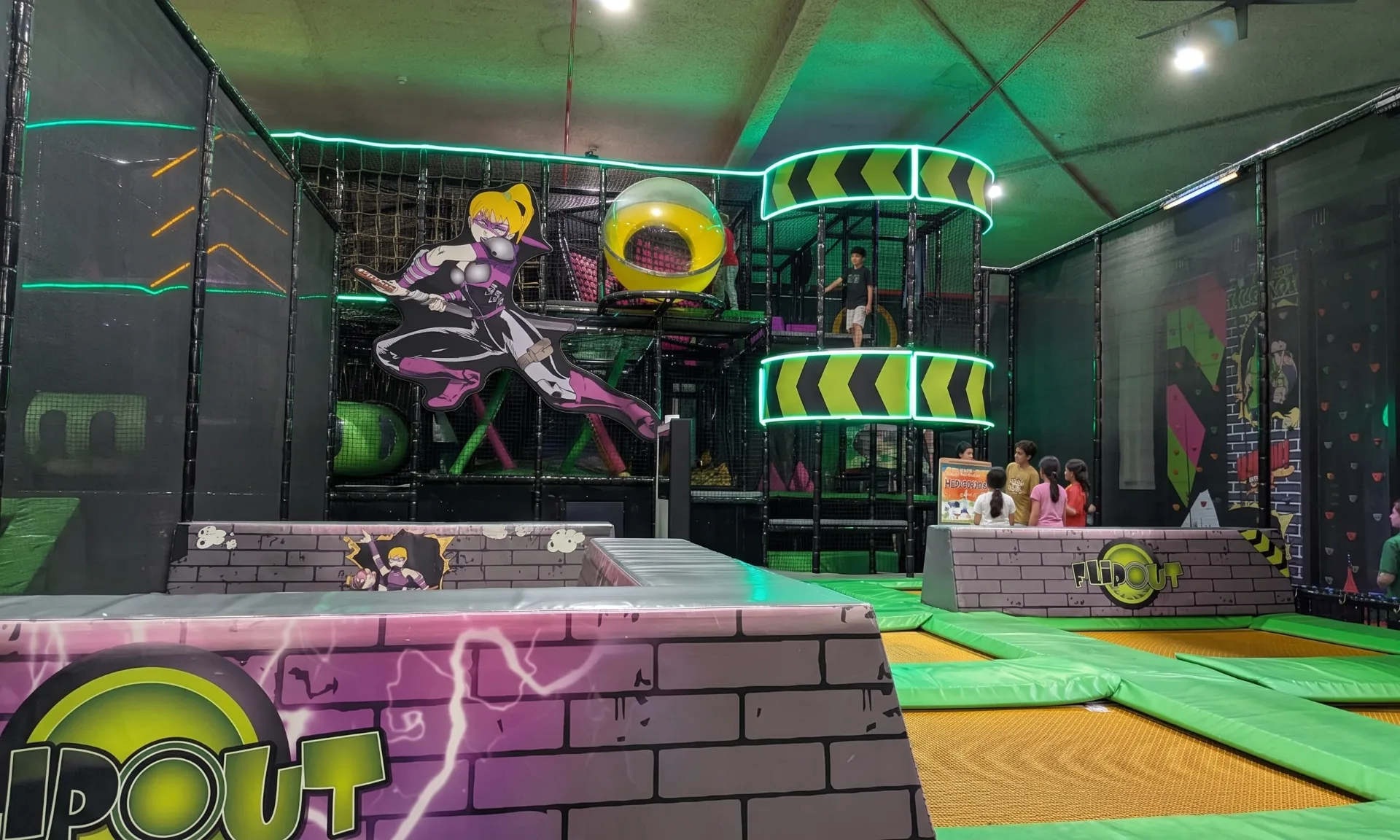 Indoor trampoline park with multi-level play structure featuring climbing nets, tunnels, and obstacle courses. Bright green and purple padding throughout with LED lighting creates an energetic atmosphere.